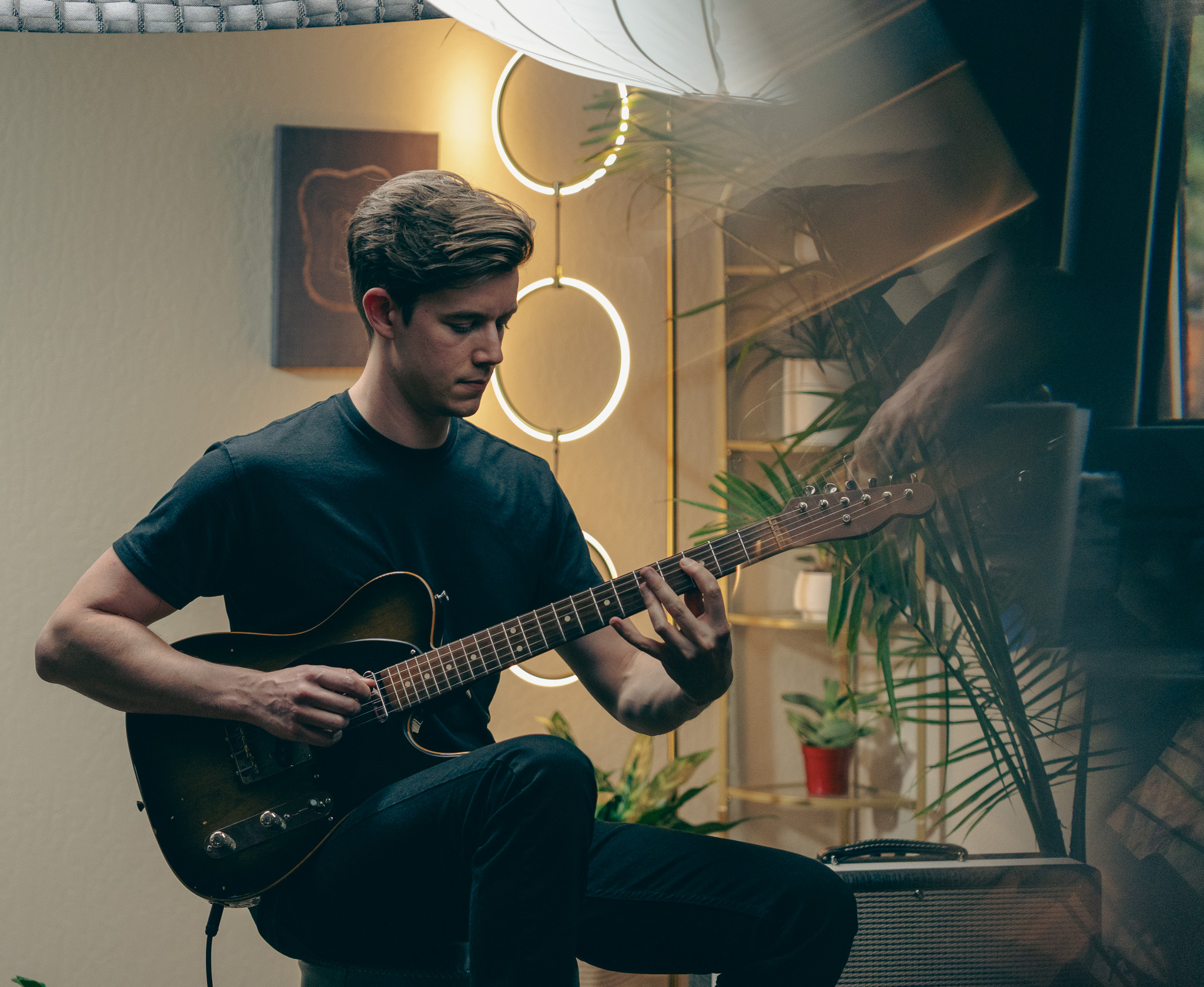 Young man playing an acoustic guitar indoors with modern lighting and plants in the background.