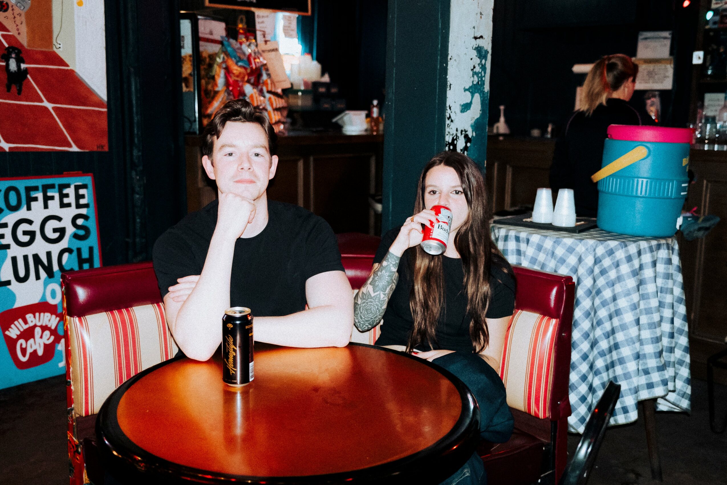 Two people sit at a round table in a cafe, one with a can and the other with a drink, in a lively setting.