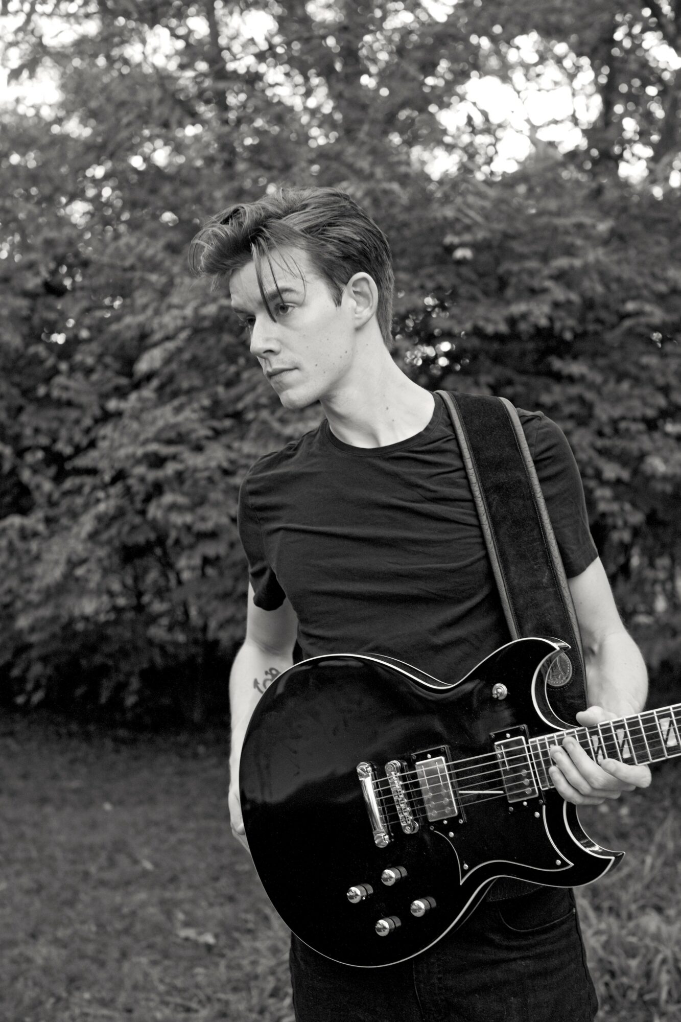 Young person with short hair holding a black electric guitar outdoors, trees in background, in black and white.