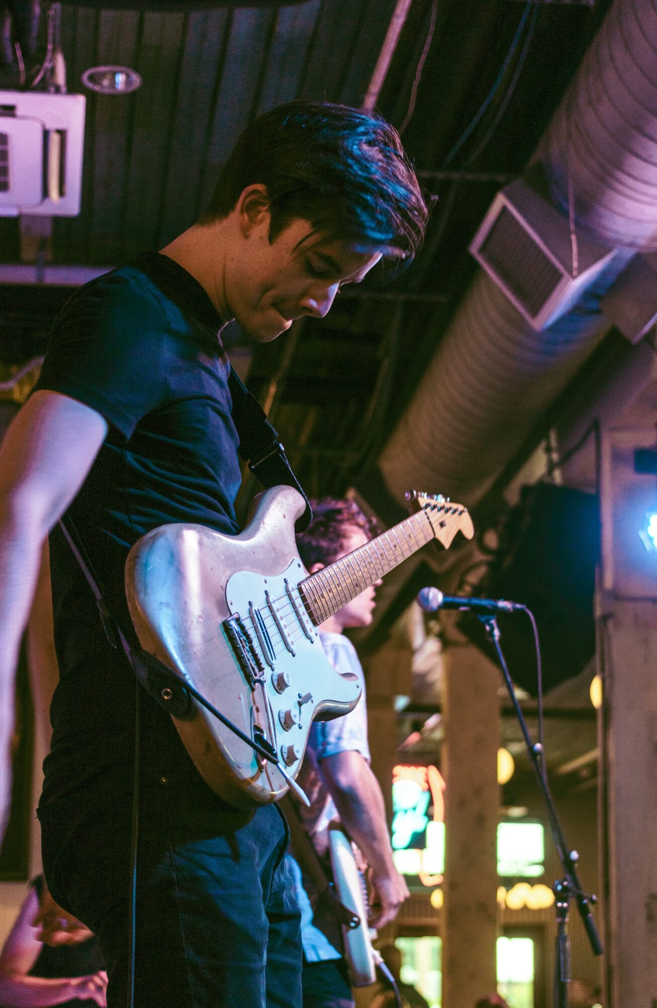 Young man playing an electric guitar on stage with dark background and stage lighting.