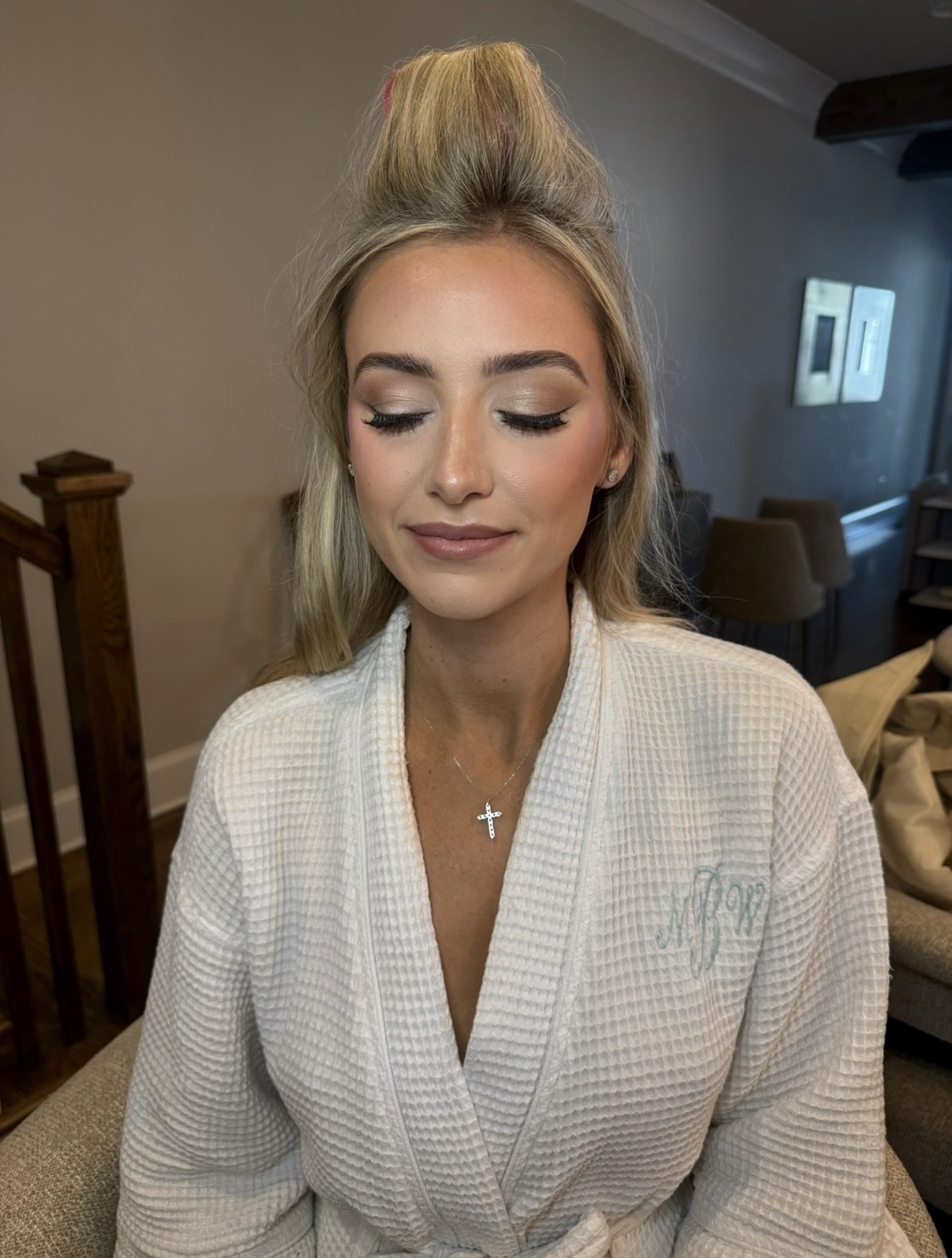 Young woman with styled hair and makeup, wearing a white robe, smiling with eyes closed in a cozy room.