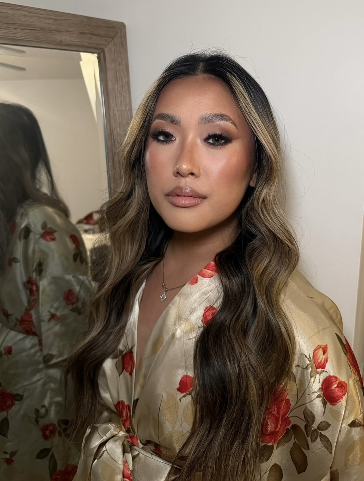 Woman with long wavy hair and makeup, wearing a floral satin robe, standing indoors near a mirror.