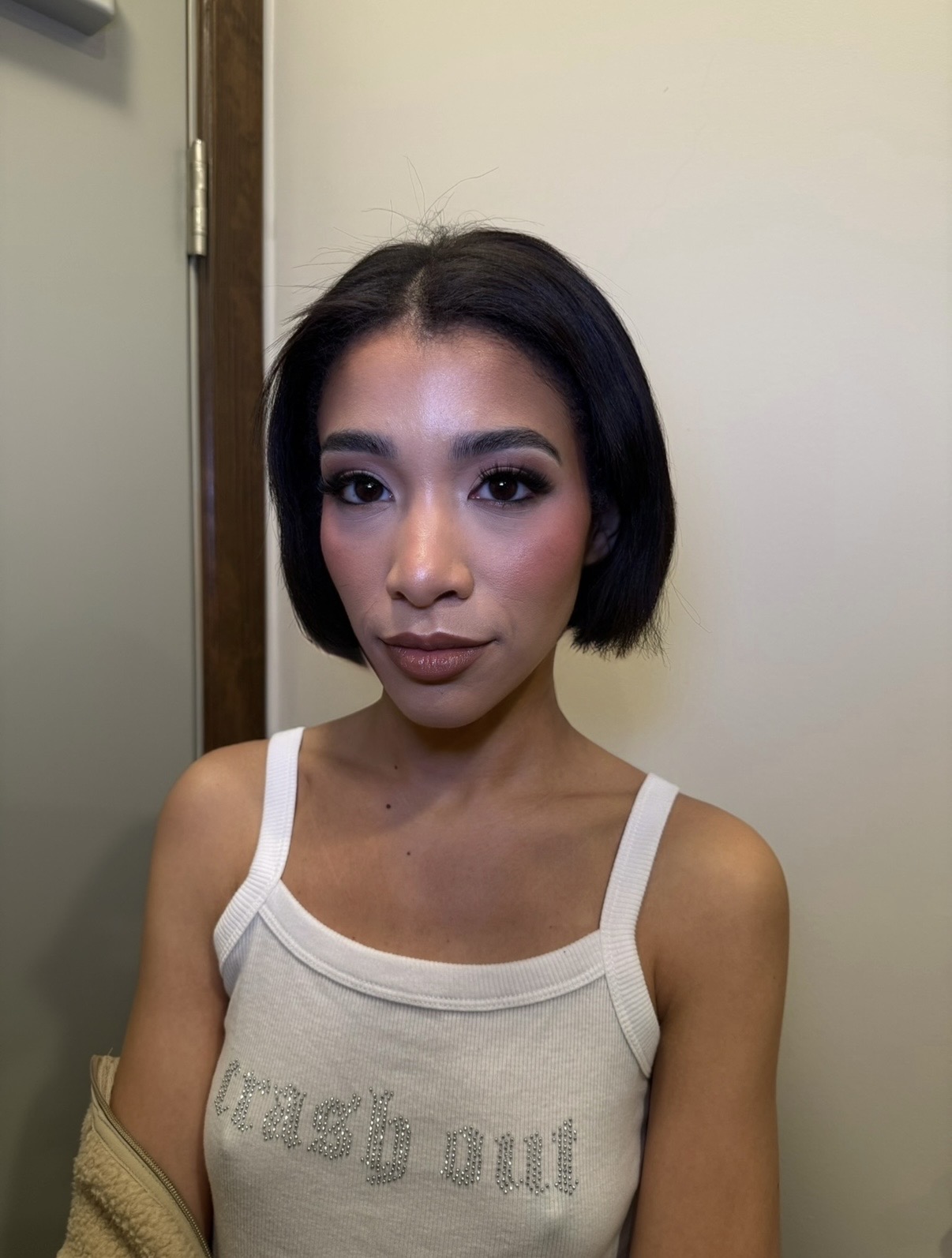 Young woman with short black hair and makeup, wearing a white tank top, standing indoors against a plain wall.