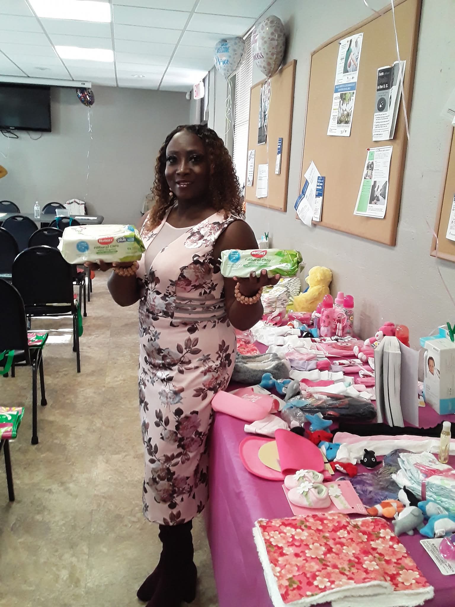 Woman standing behind table with various colorful handmade items, holding two decorated cakes, in a room with bulletin boards.