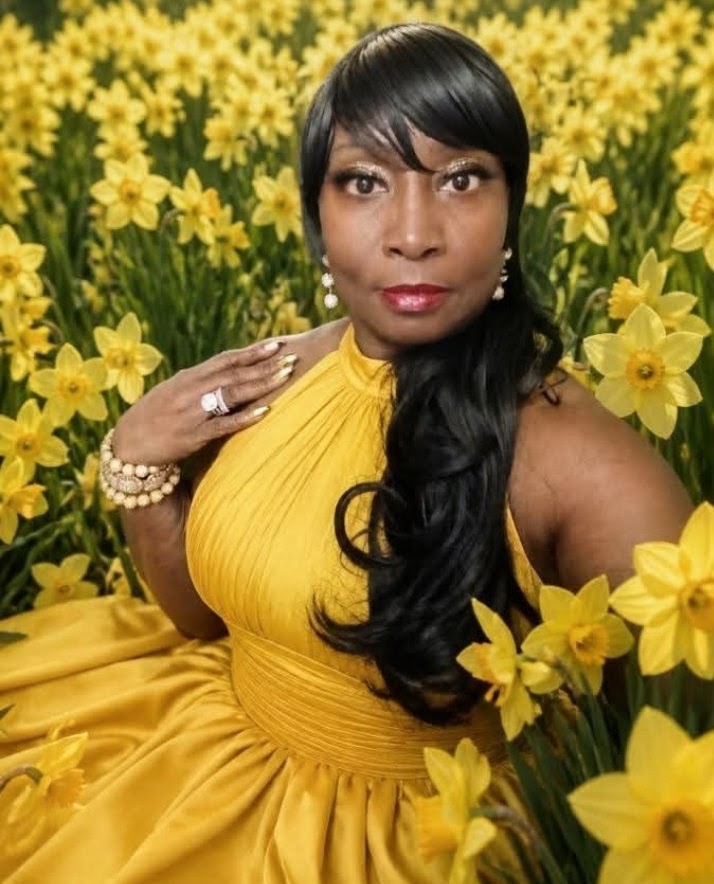 Woman in a yellow dress with long black hair, surrounded by yellow flowers, looking at the camera.