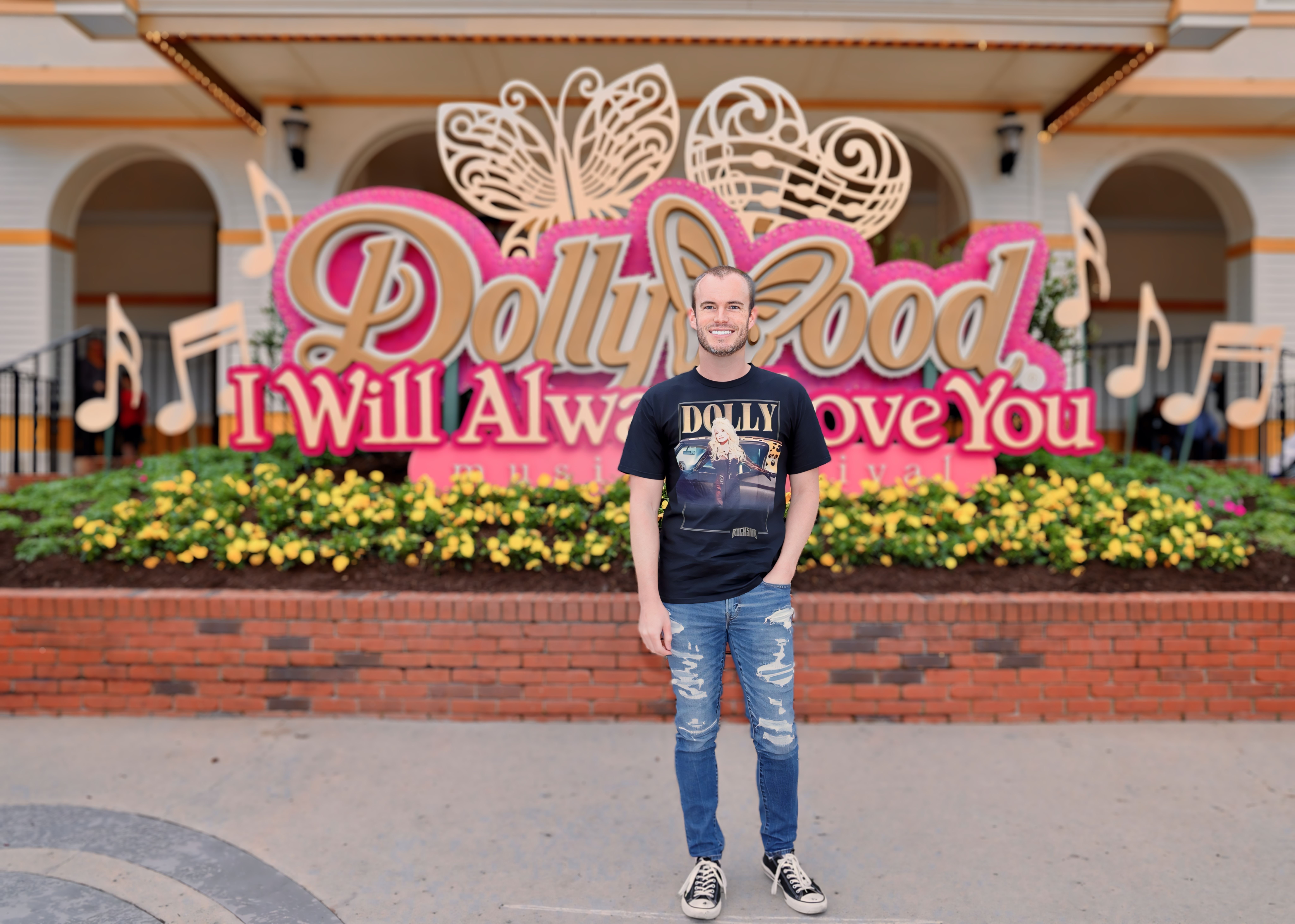 Person standing in front of a Dolly Parton-themed display with flowers and musical notes, smiling, wearing a black T-shirt and jeans.