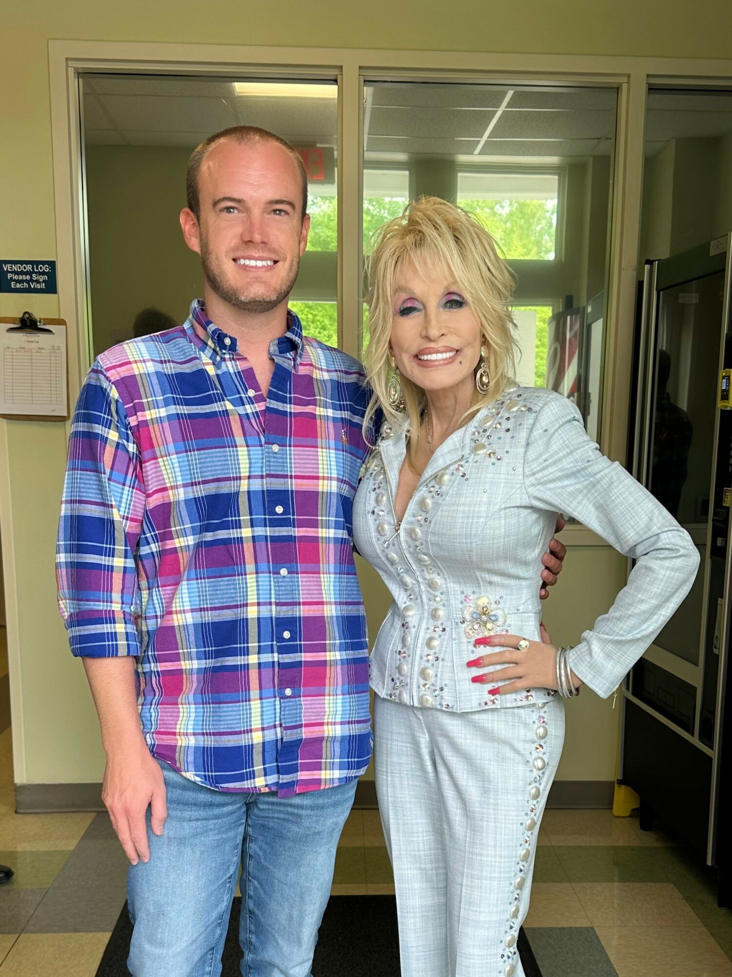 Two people standing indoors, smiling, with a window and office equipment in the background.