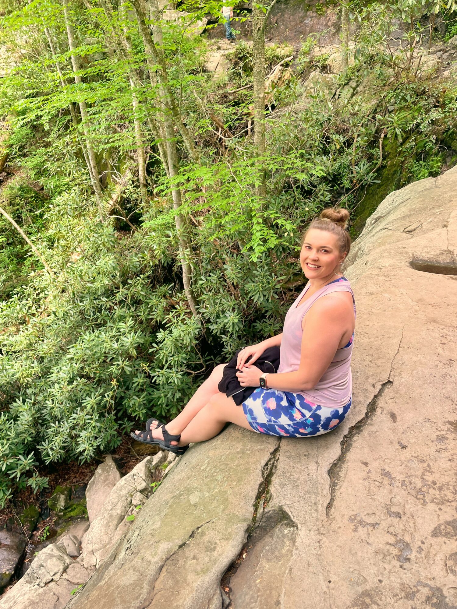 Young woman sitting on a large rock outdoors, surrounded by green trees and foliage, smiling at the camera.