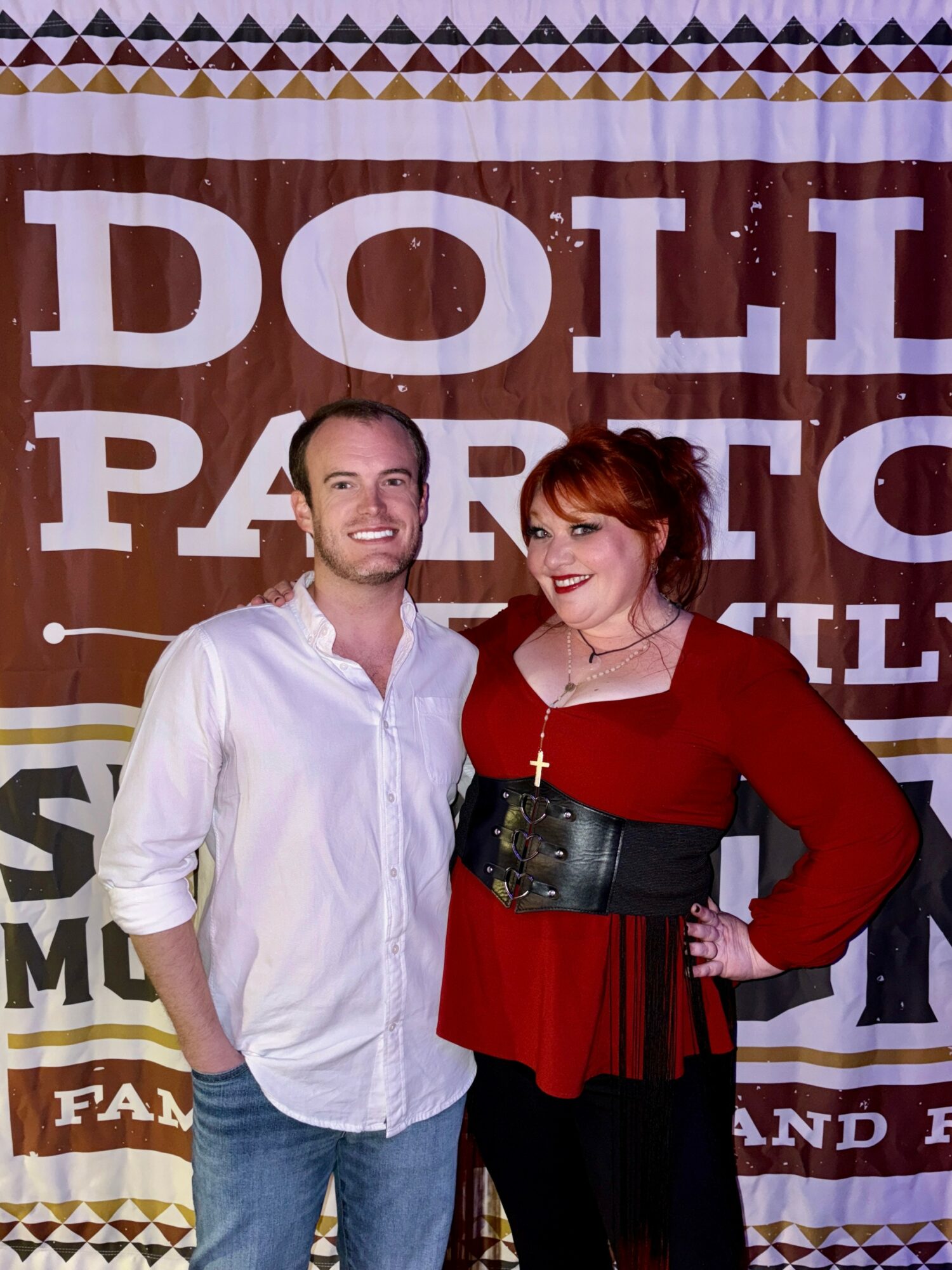 Two people standing in front of a large banner, smiling, with one person wearing a white shirt and the other in a red top with a black belt.