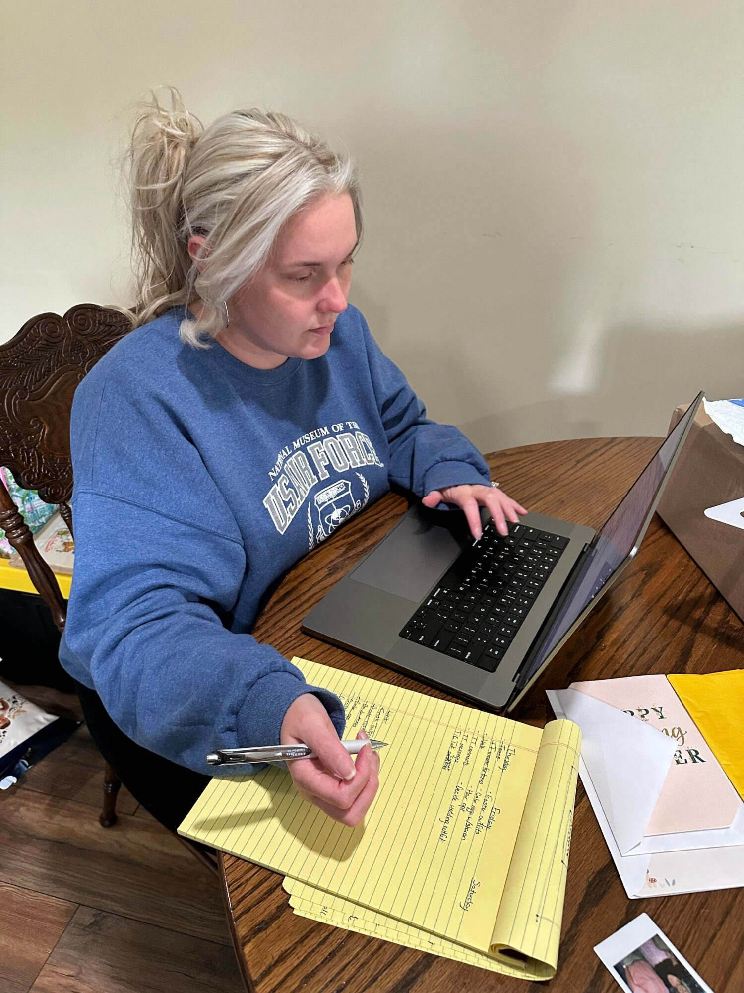 Woman with blonde hair in a blue sweatshirt writing on a yellow notepad at a table with a laptop.