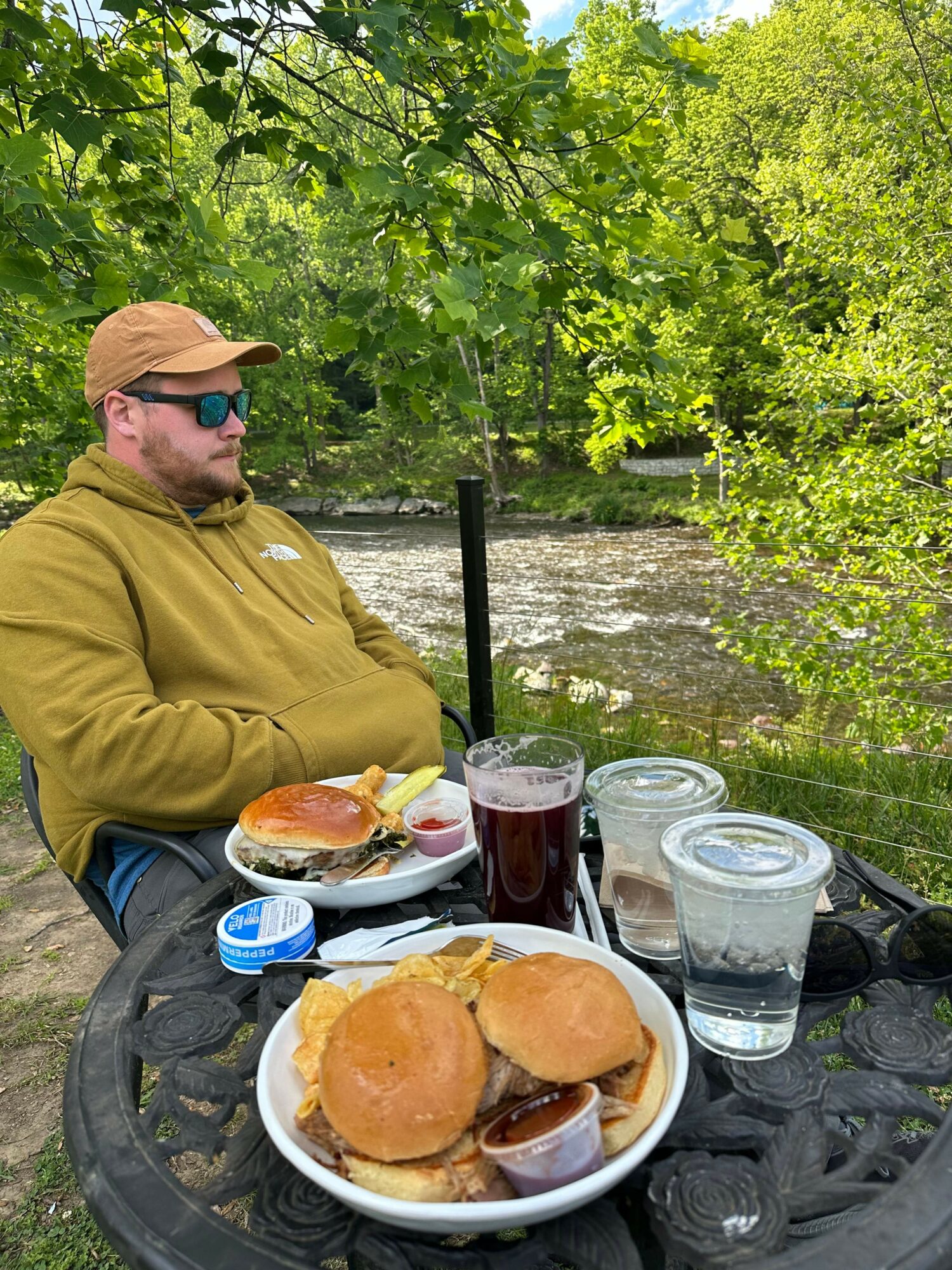 Person wearing a yellow jacket and cap sitting outdoors at a table with food and drinks, surrounded by green trees.