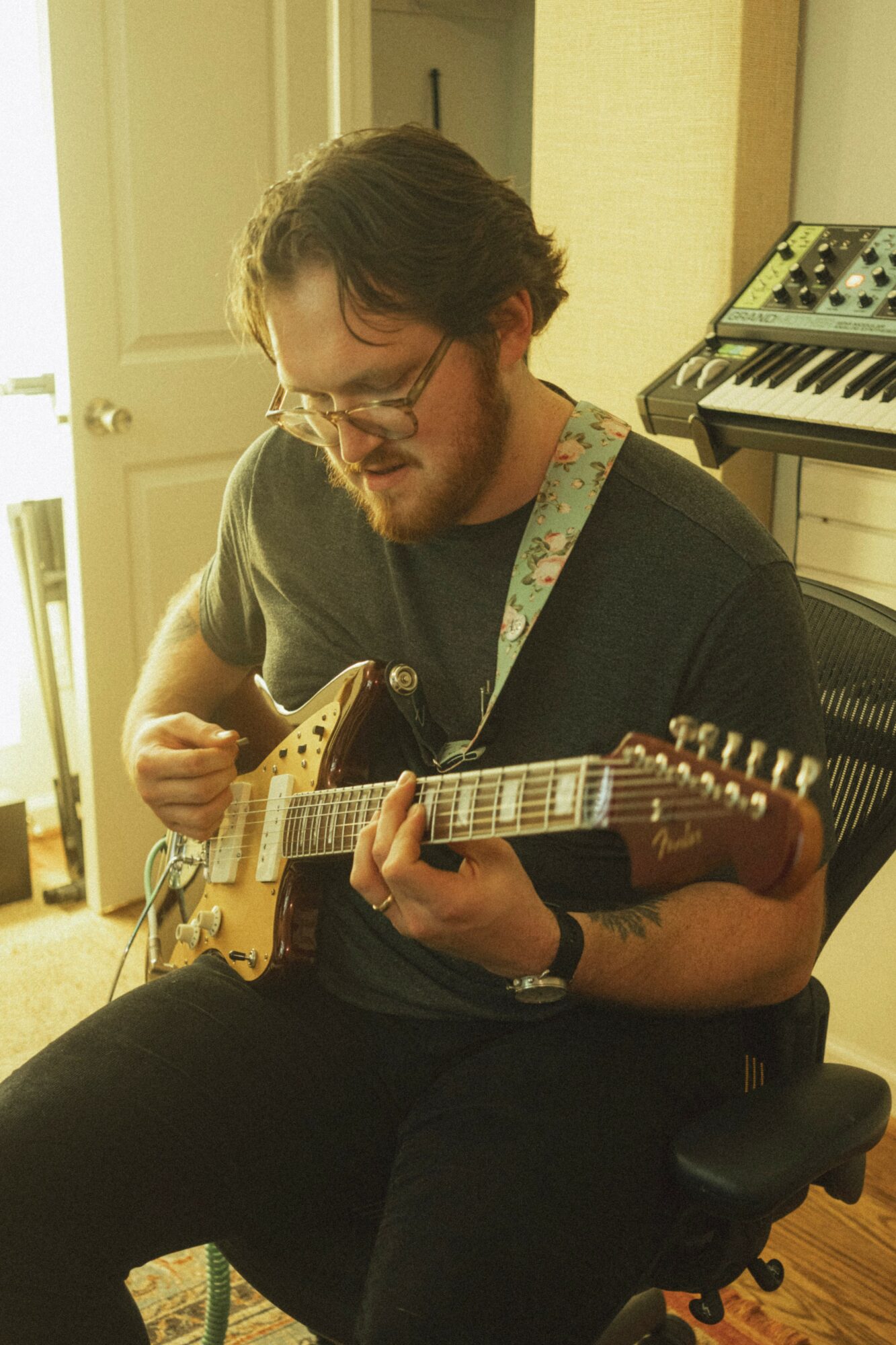 Person with glasses and beard playing an electric guitar in a room with musical equipment.