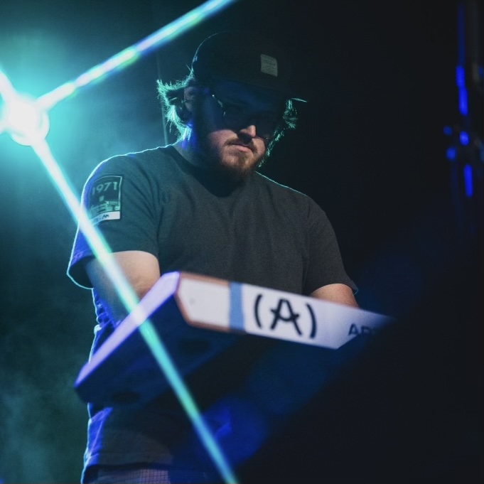 Person wearing headphones and sunglasses, playing a keyboard on stage with bright lights behind them.