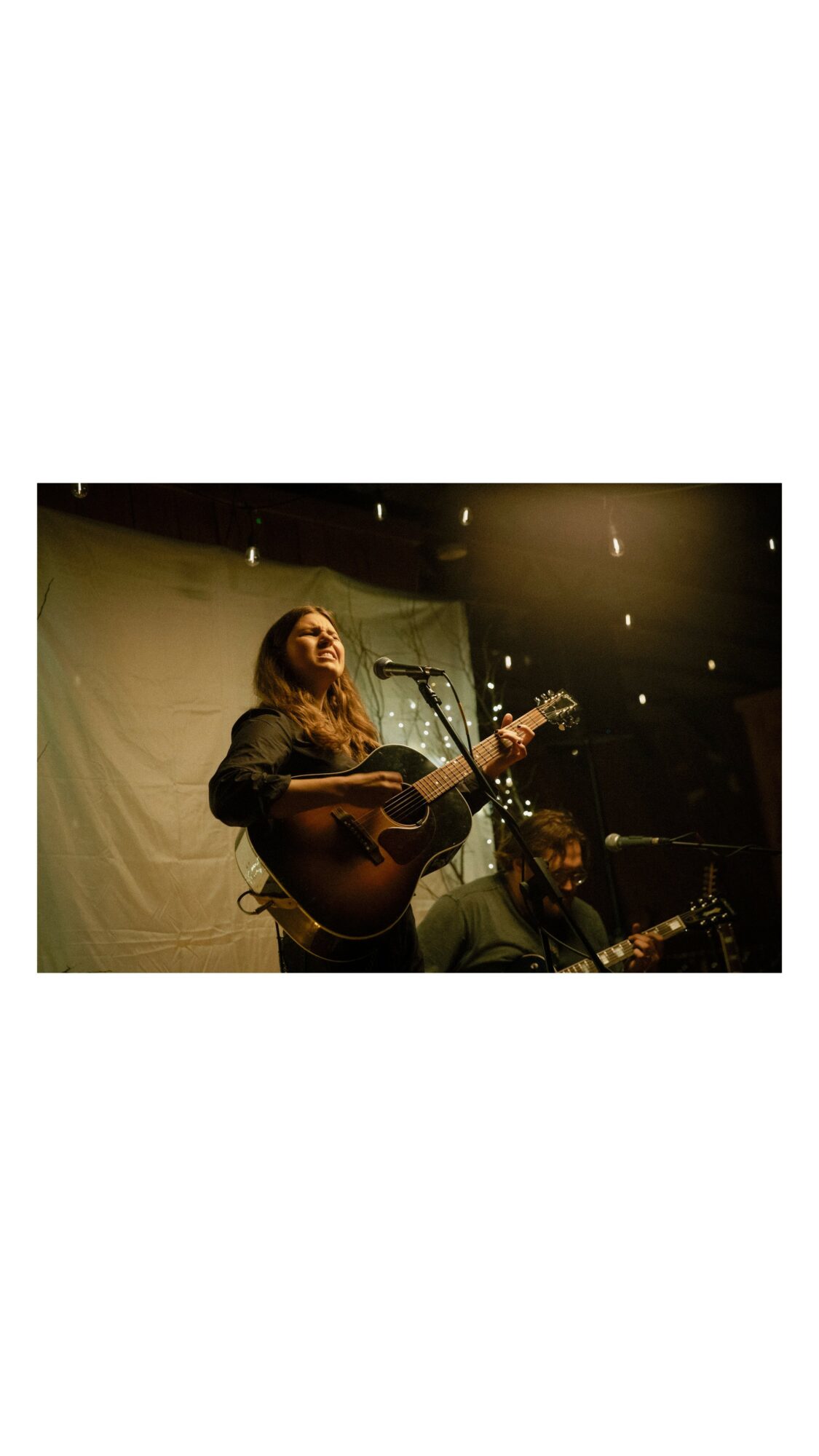 Person singing and playing guitar on stage with another person in the background, dim lighting, dark backdrop.