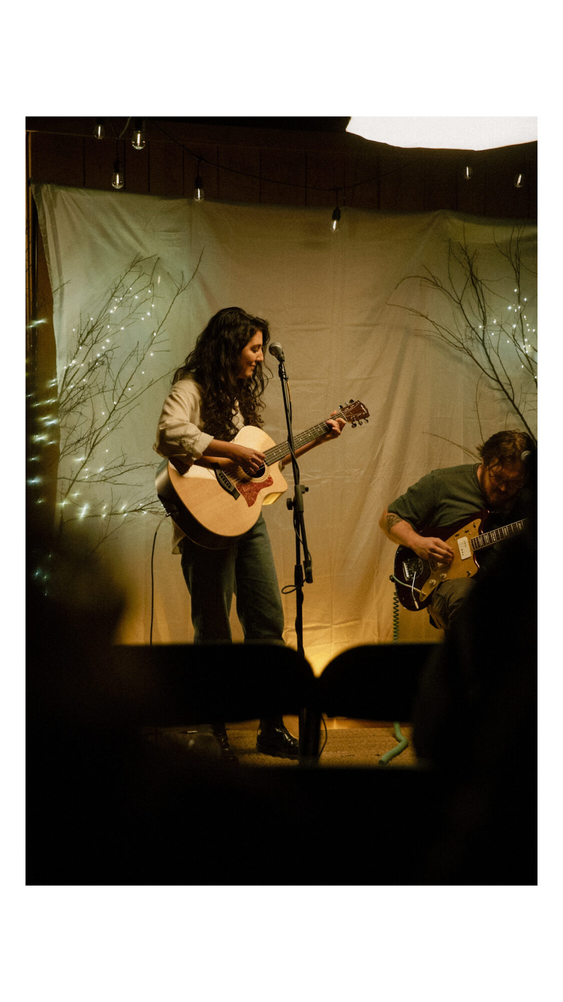 Two musicians perform on stage, one playing guitar and singing, the other playing guitar, with a decorated backdrop.