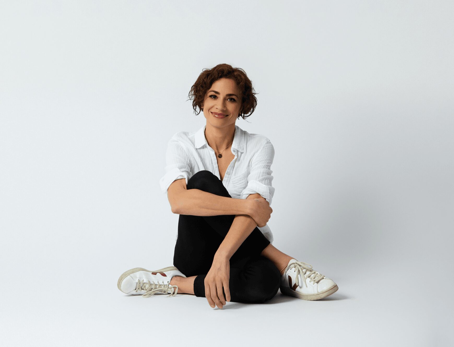 Woman sitting cross-legged on the floor, smiling, wearing a white shirt, black pants, and white sneakers, against a plain background.