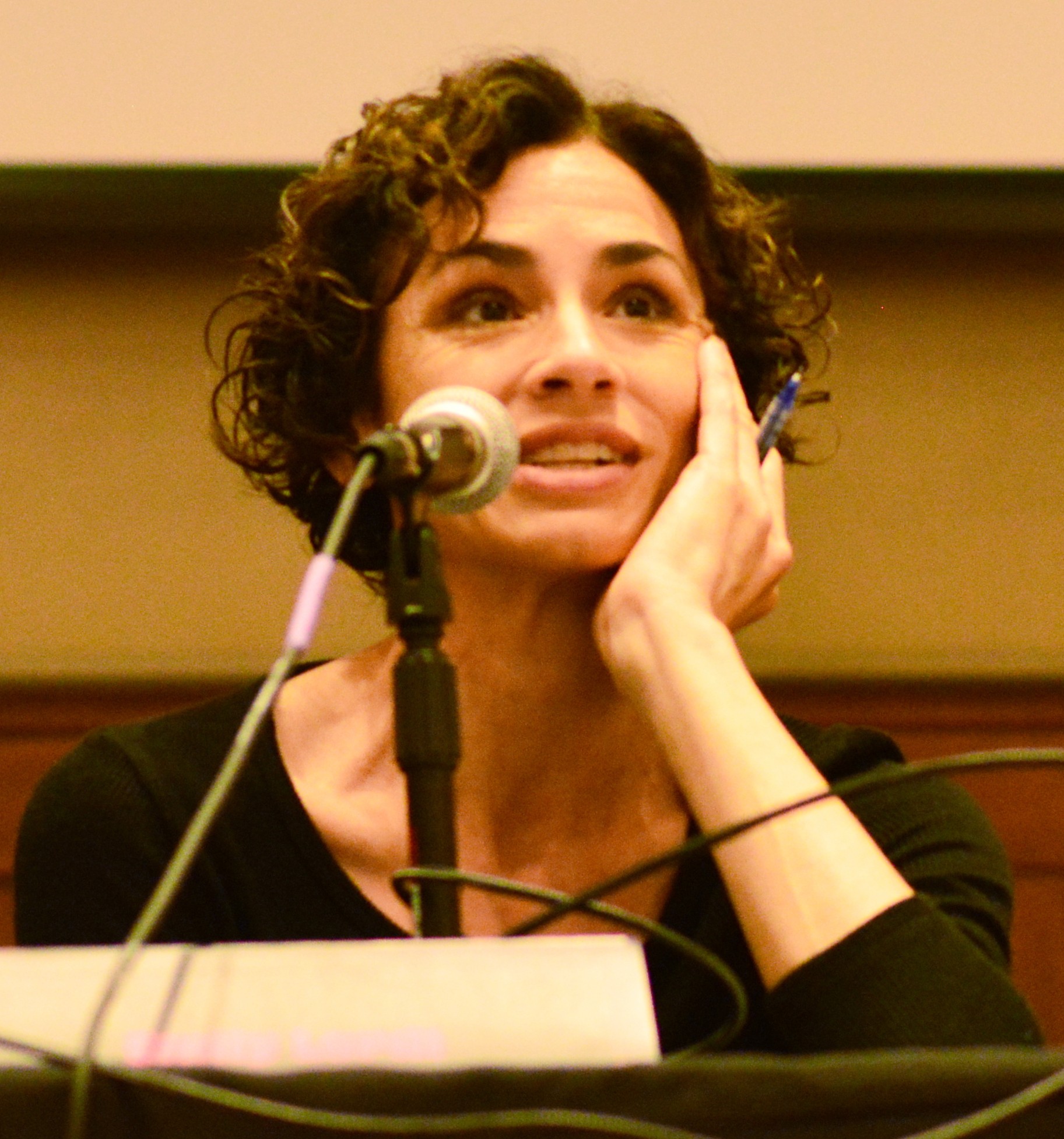 Woman with curly hair speaking into a microphone, resting her face on her hand, in a panel or conference setting.