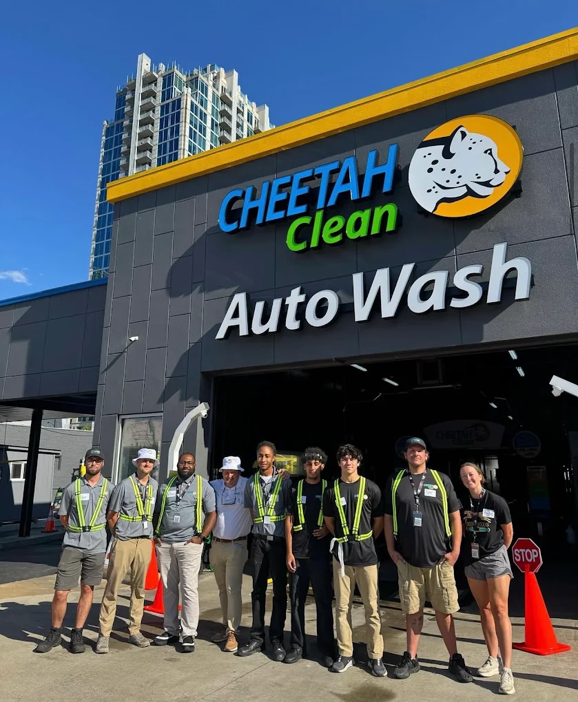 Group of people standing outside a car wash with a sign reading 'Cheetah Clean Auto Wash' and a tall building in the background.