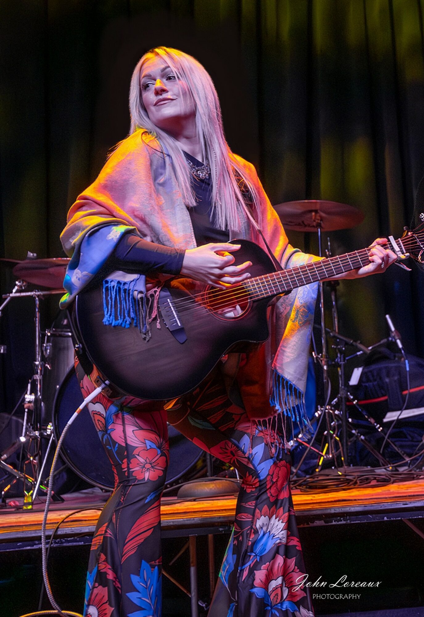 Woman with long blonde hair holding a guitar on stage with a dark background.
