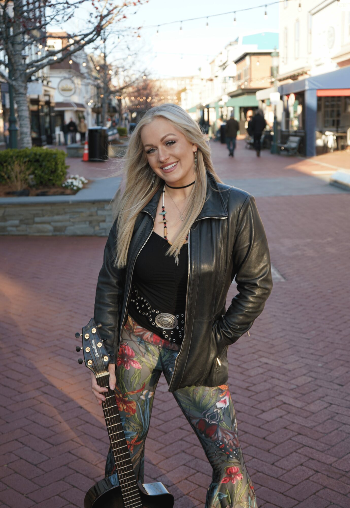 Woman with long blonde hair smiling, holding a guitar, wearing a black leather jacket and colorful floral pants, on a city street.