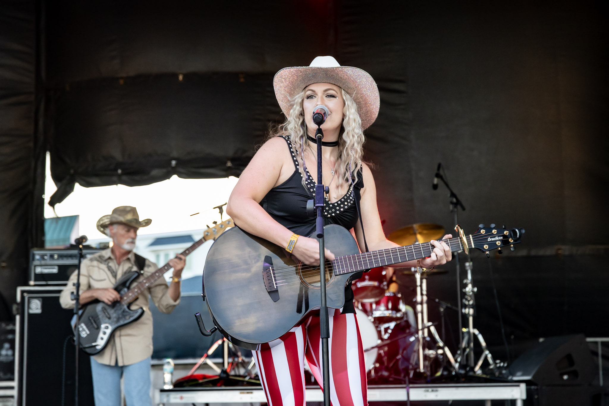 Female musician with a guitar and hat performing on stage with band members in background.
