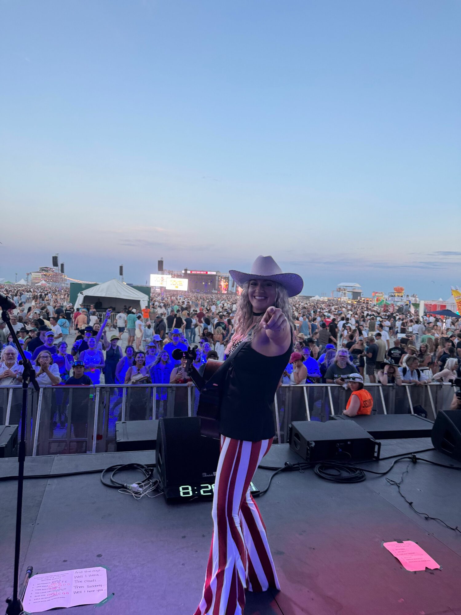 Person with long hair and hat standing on stage with crowd in background, performing at outdoor event during sunset.