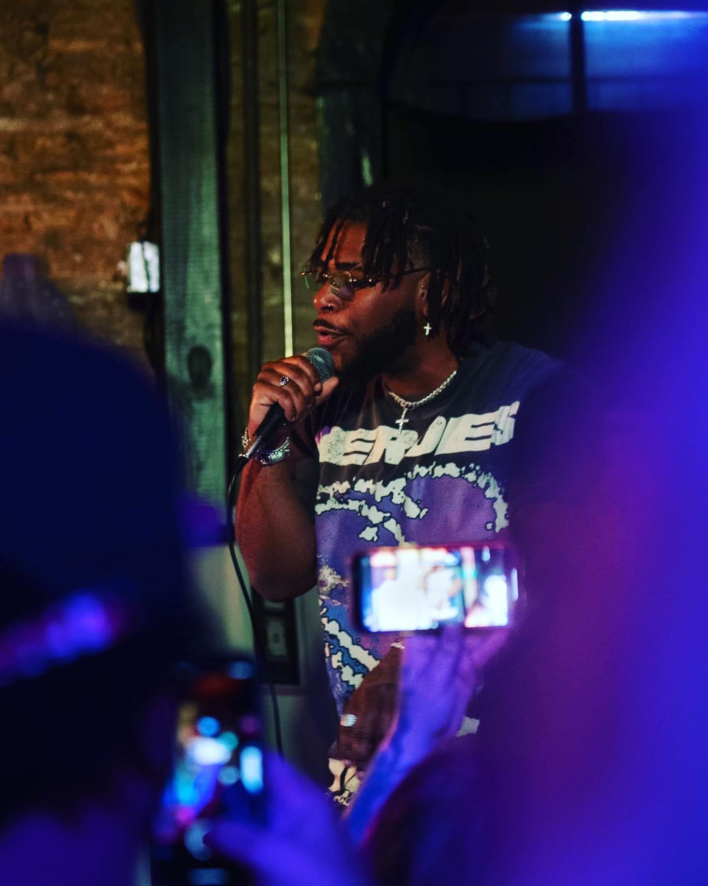 Person with dreadlocks holding a microphone, speaking or singing in a dimly lit setting with colorful lights.