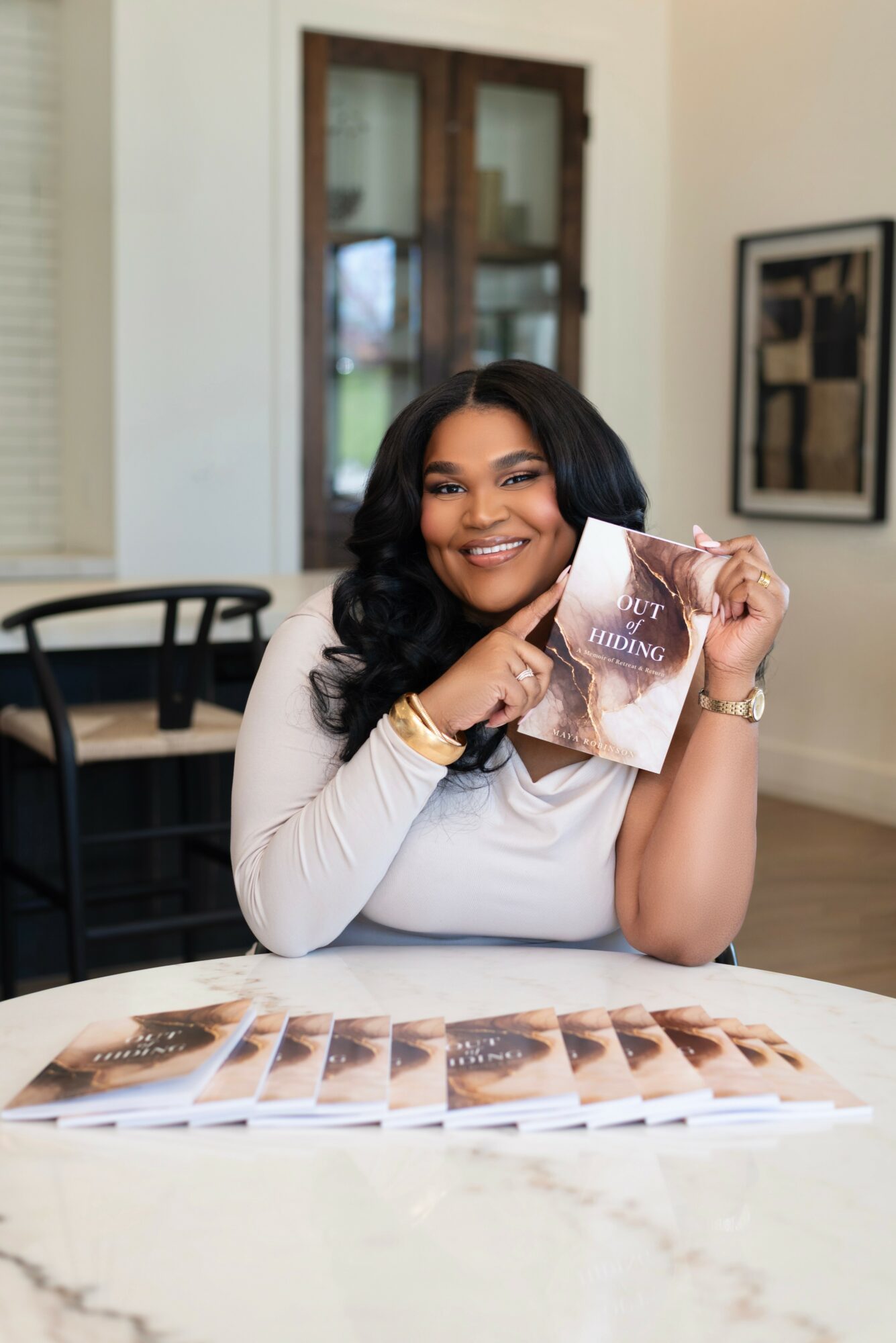 Smiling woman with dark hair holding a book, sitting at a table with brochures, in a room with artwork and a window.