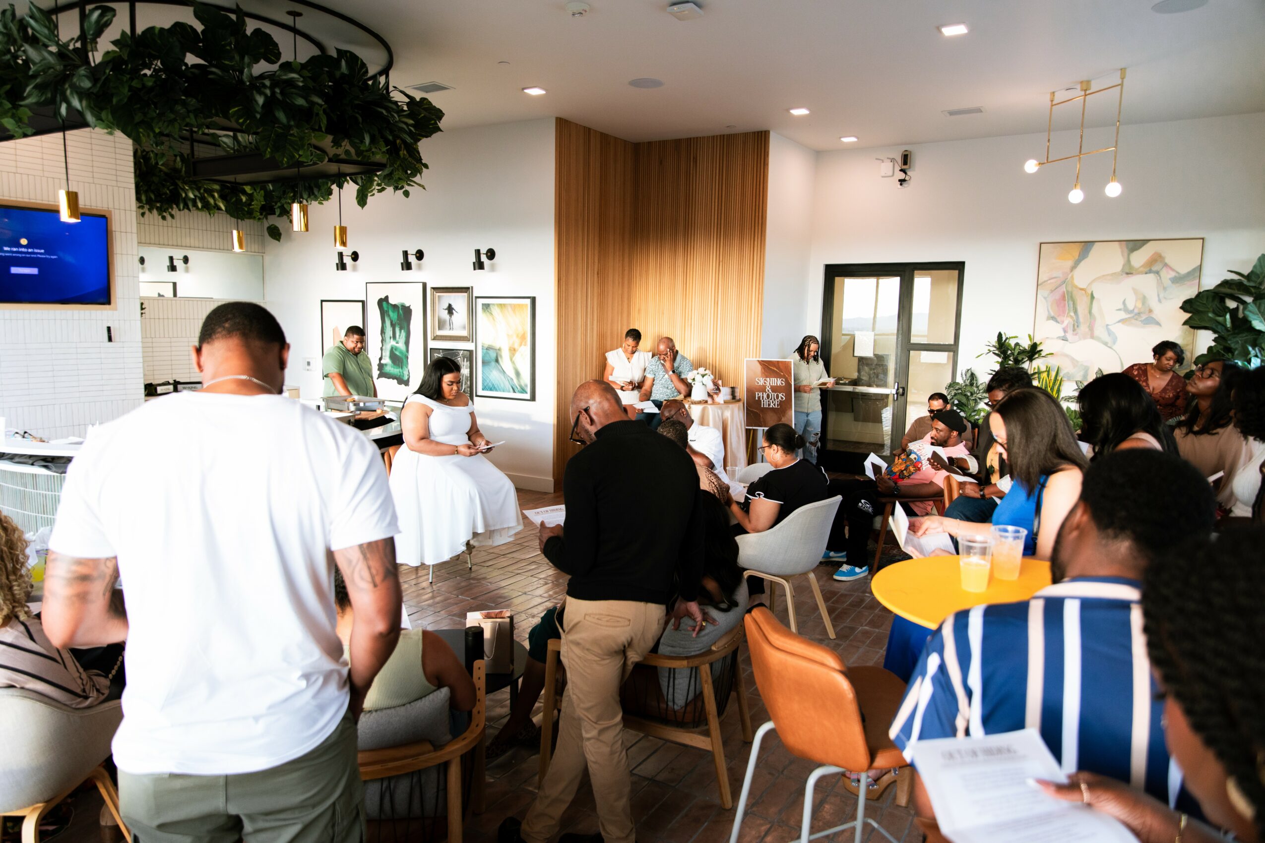 People gathered in a room with a wooden wall, some sitting at tables, others standing, watching a performance or presentation.