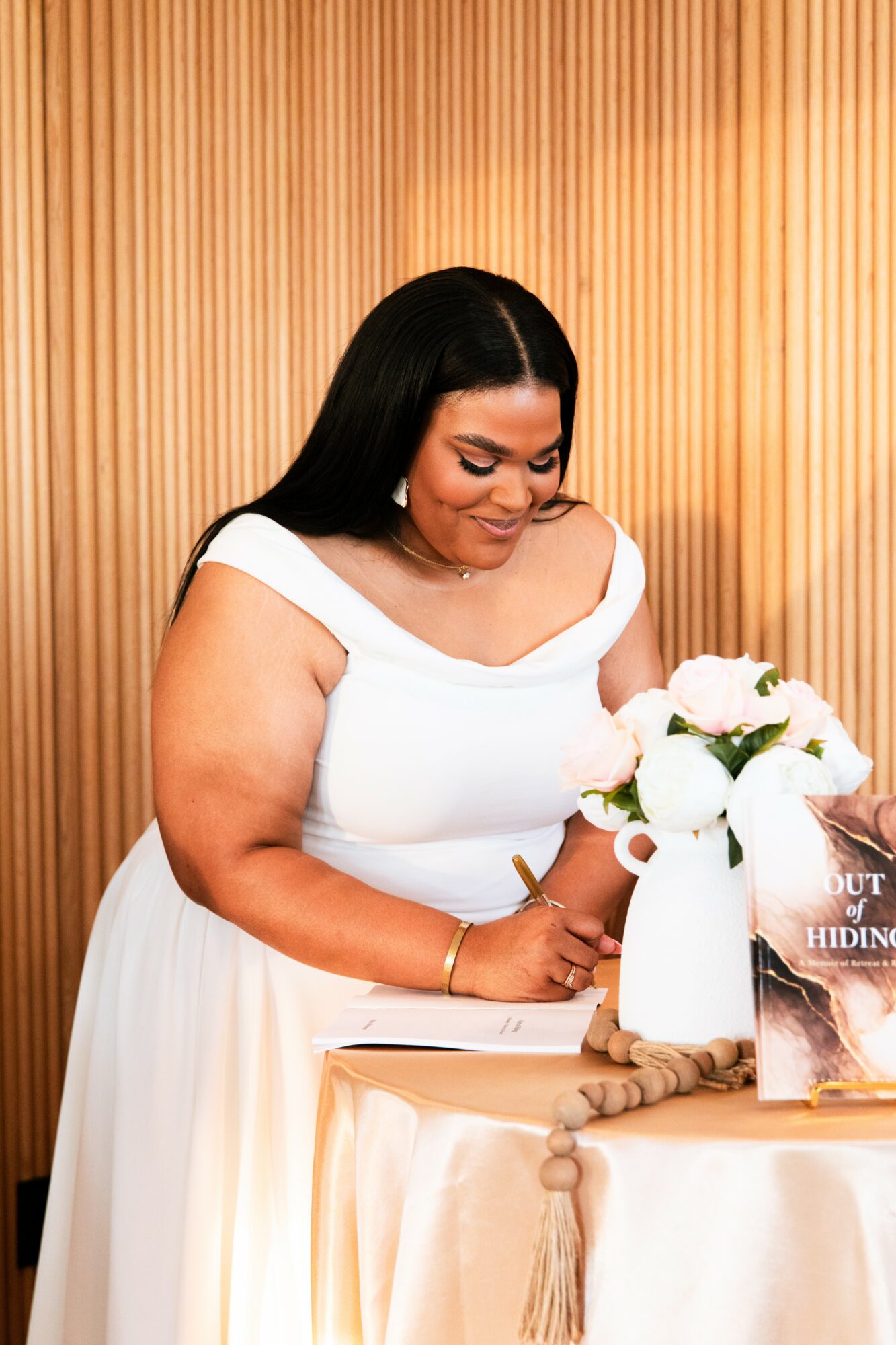 Woman in white dress with long dark hair, standing at a table with a white flower arrangement, wooden background