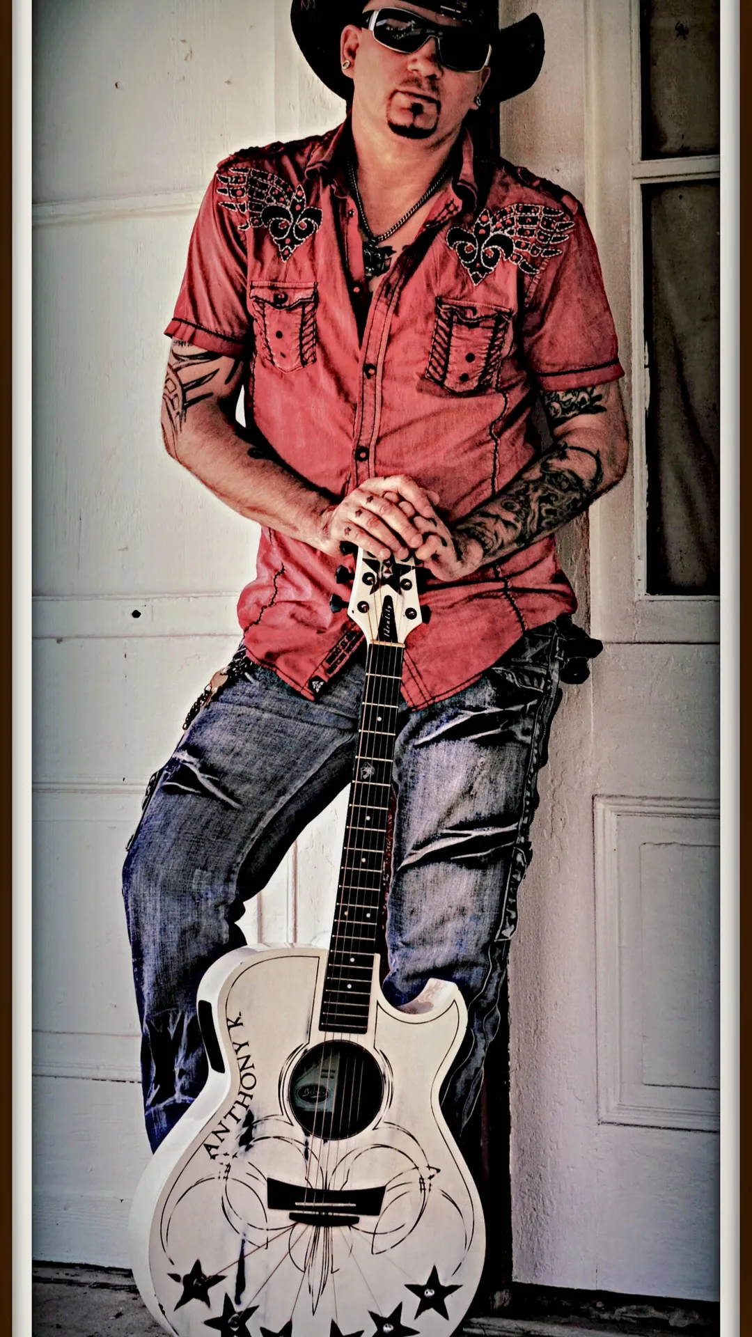 Person wearing a hat and sunglasses, holding a guitar, standing indoors near a door, dressed in a red shirt and jeans.
