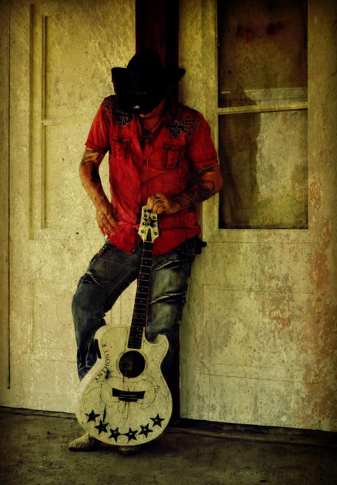Person wearing a hat and red shirt, holding a decorated guitar, standing against a wall with a window.