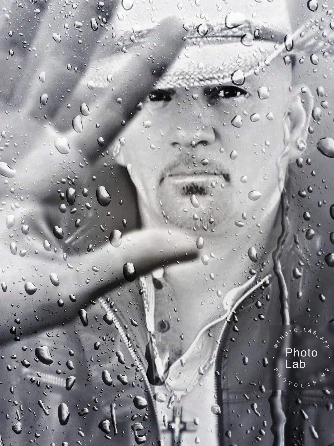 Person wearing a hat and hoodie, looking through a rain-covered glass, with a serious expression, hand raised close to the glass.