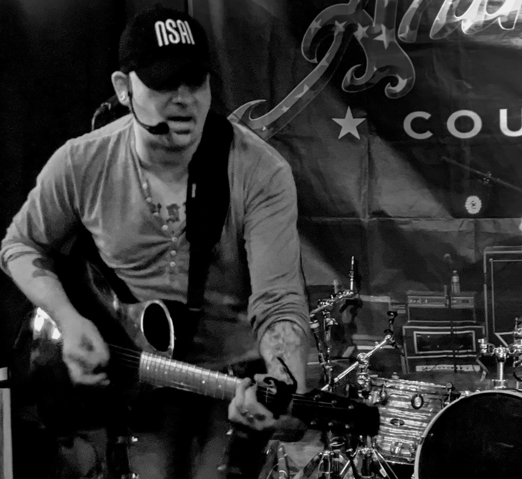 Person playing guitar on stage with musical equipment in background, wearing a cap and casual clothing.