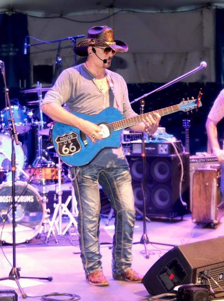 Person wearing a cowboy hat playing a blue guitar on stage with drums and equipment in background.