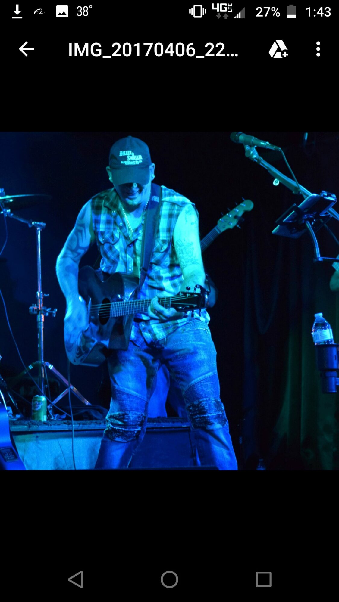 Person playing guitar on stage with microphone and water bottle nearby, dark background, blue lighting.
