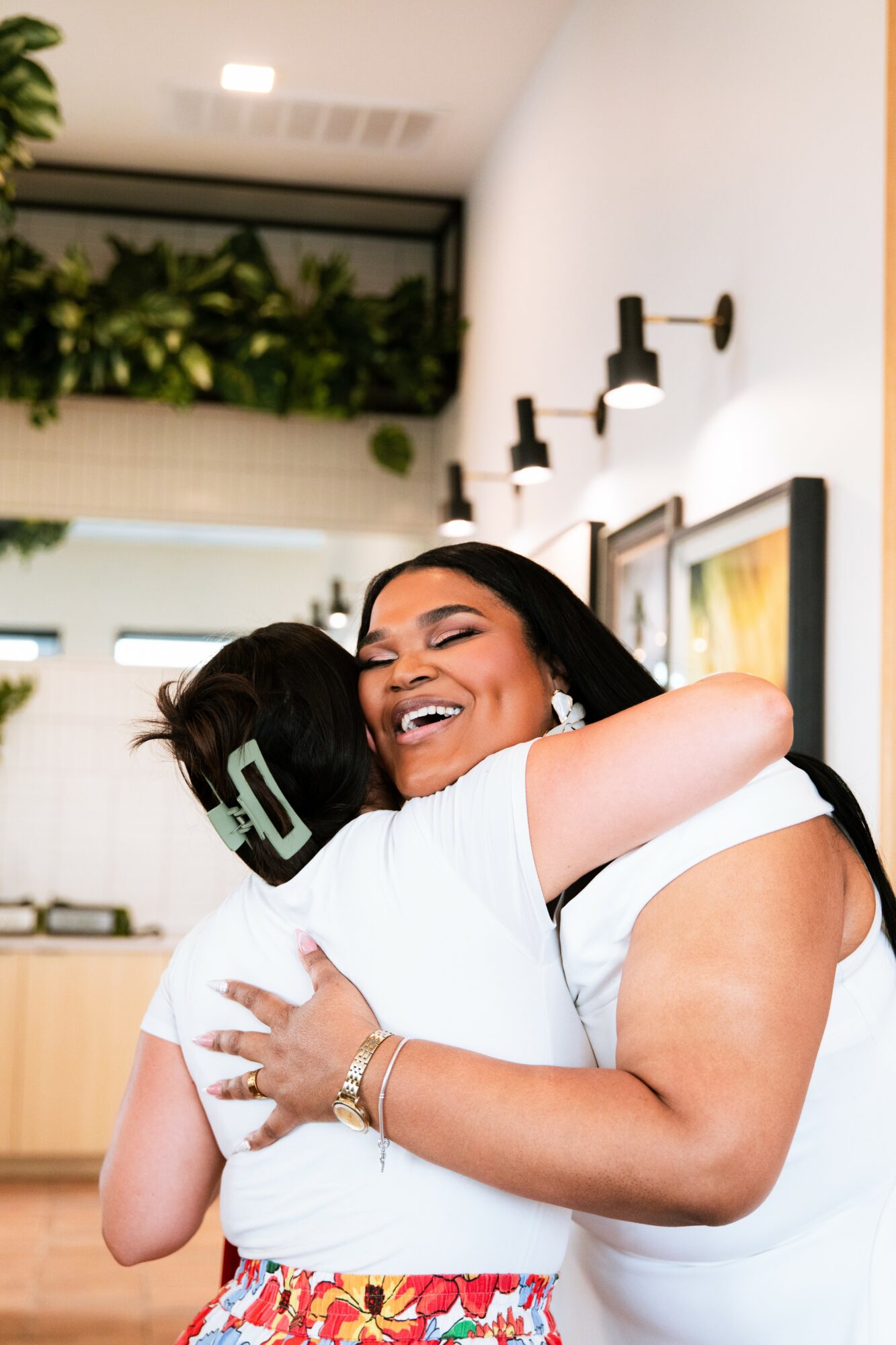 Two women hugging and smiling in a modern kitchen with wall-mounted lights and greenery above.