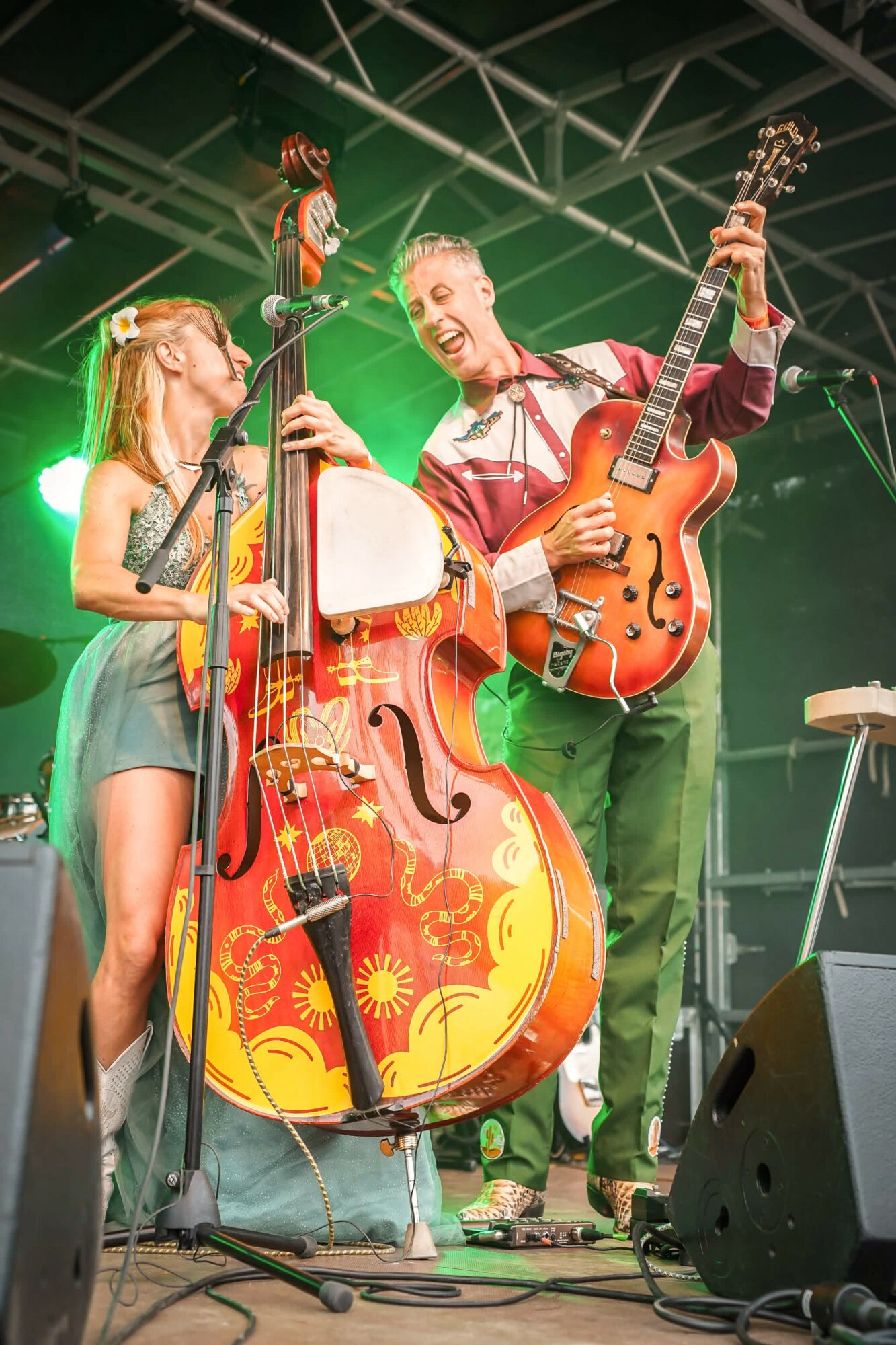 Two musicians, a woman and a man, perform on stage with guitars and a large decorated double bass, under stage lights.