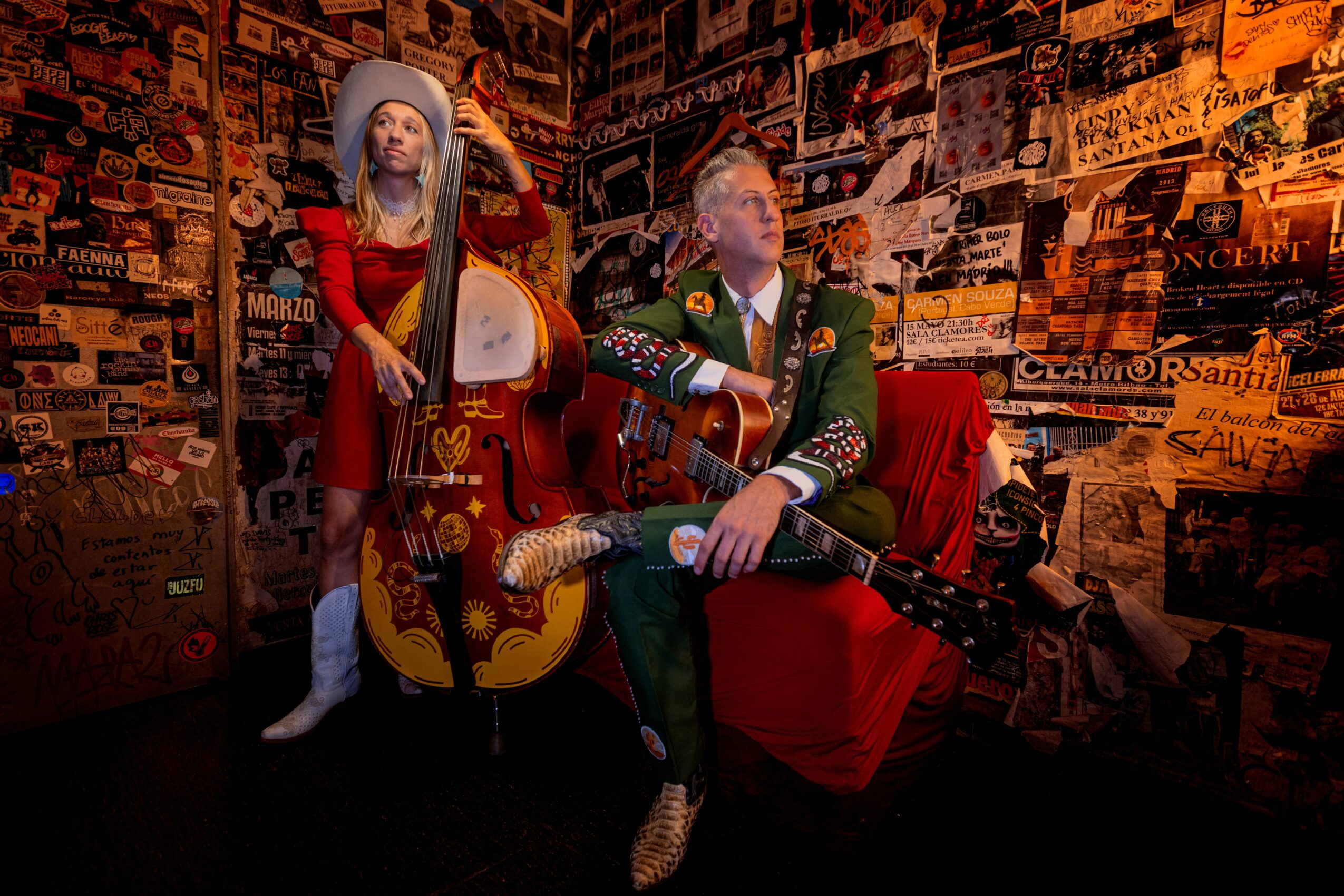 Two people with guitars sit in a room with a collage of posters on the wall behind them.
