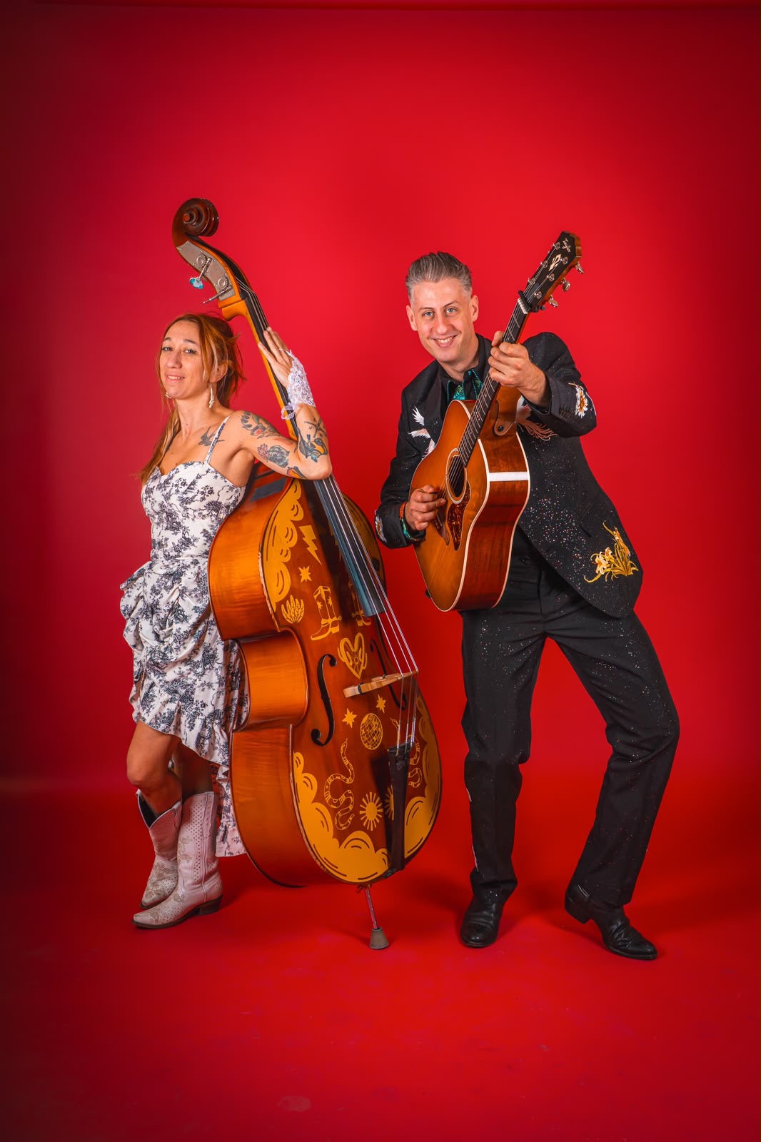 Two musicians, a woman with a double bass and a man with an acoustic guitar, stand against a red background.