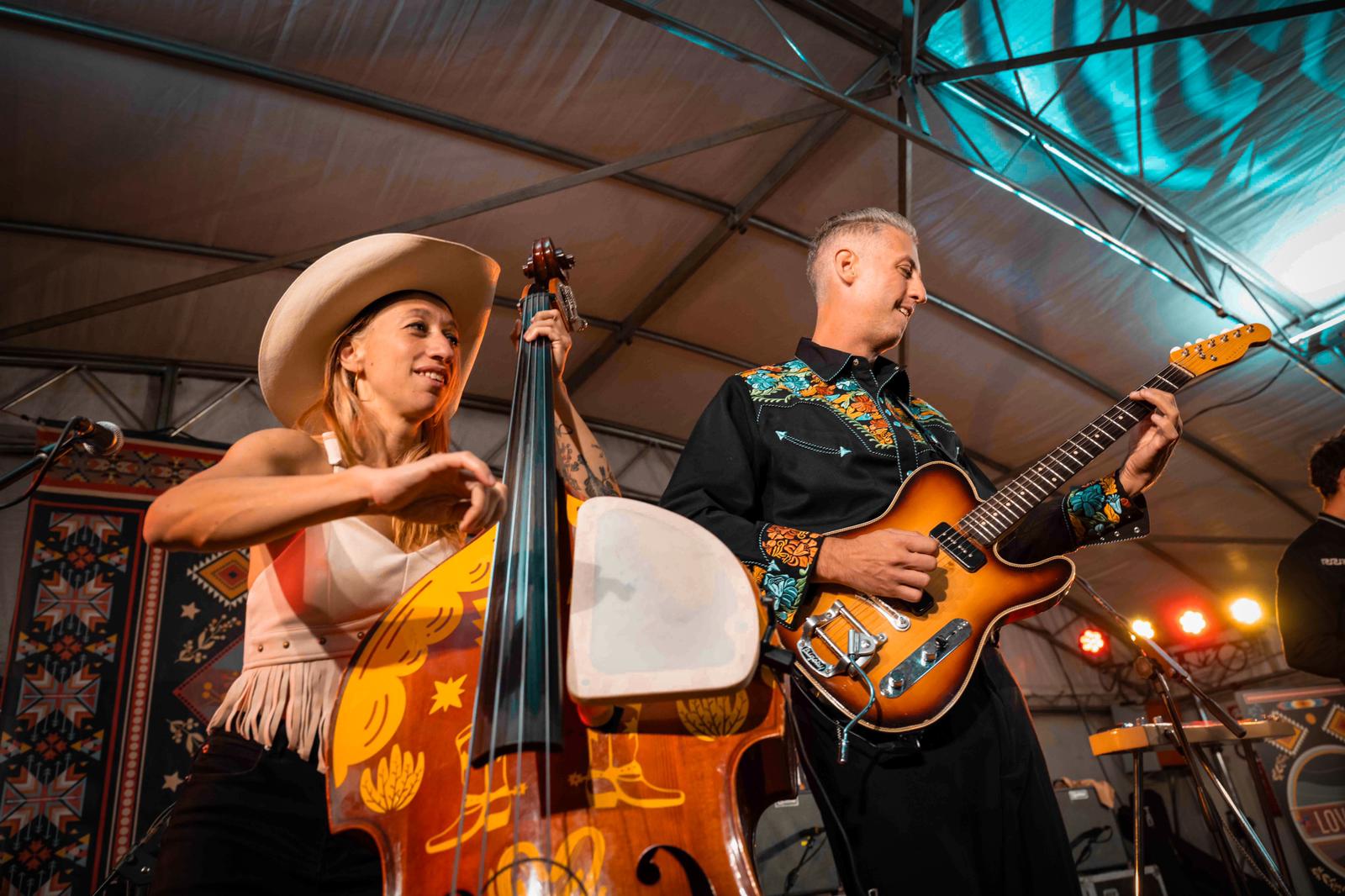 Two musicians performing on stage, one playing a double bass and the other playing an electric guitar, under a tent roof.