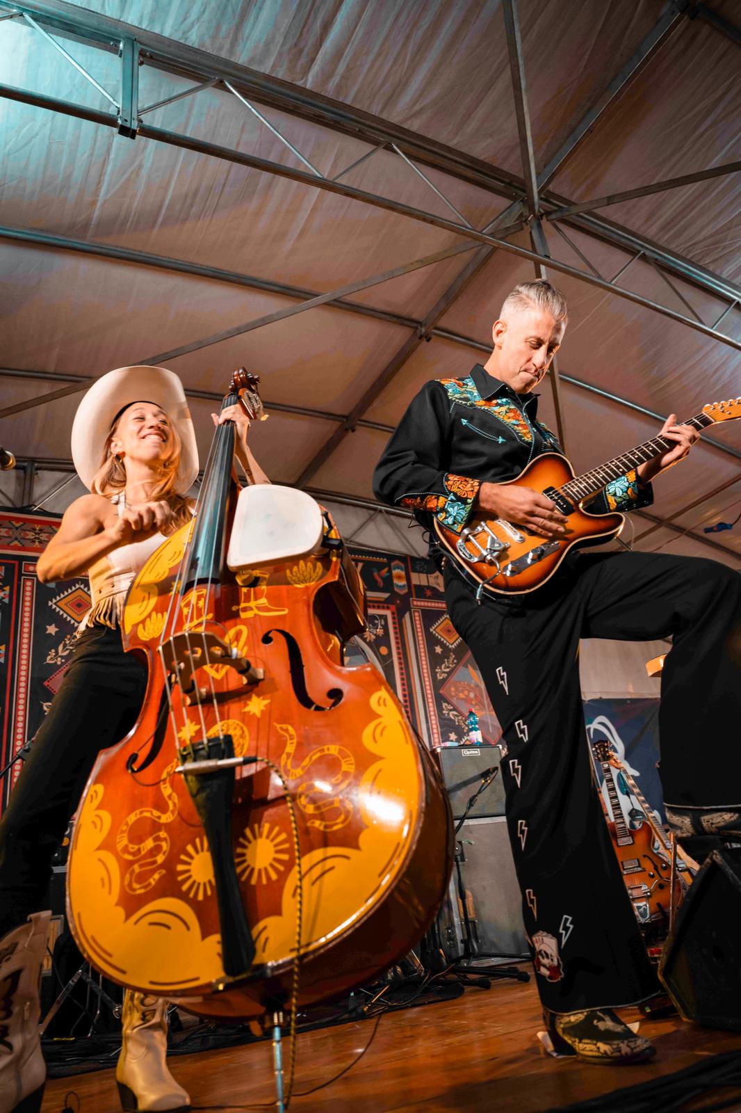 Two musicians perform on stage, one playing a double bass with floral patterns and the other playing an electric guitar, under a tent roof.