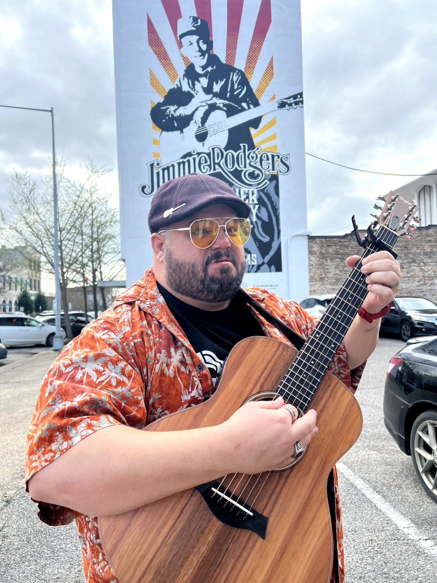 Man with glasses and a hat playing a guitar outdoors, with a Jimmie Rodgers poster in the background.