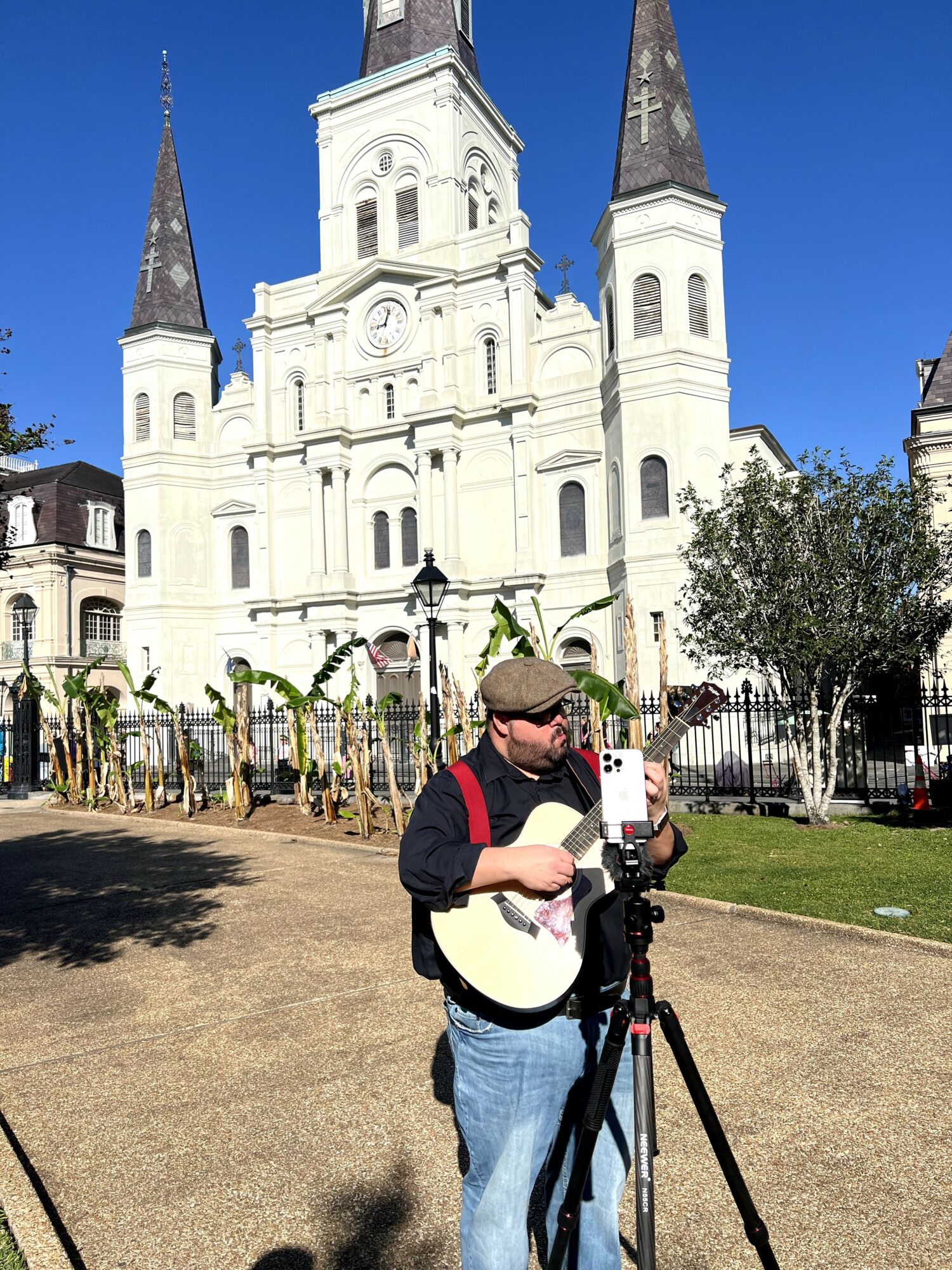 Person with a guitar and camera tripod in front of a white church with four spires, trees, and a fence.