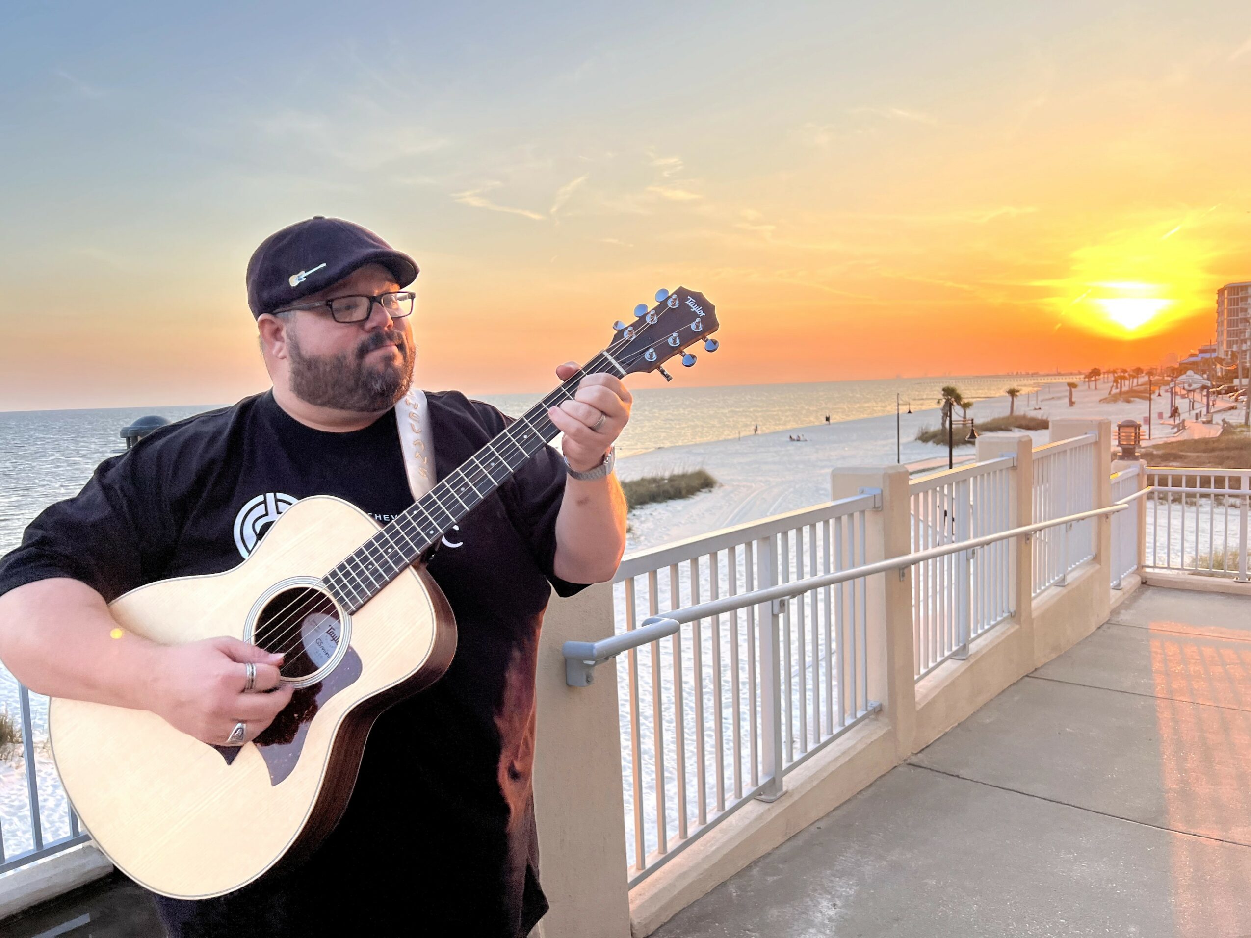 Man playing guitar on a seaside promenade during sunset, wearing a black cap and glasses, with a railing and ocean in background.