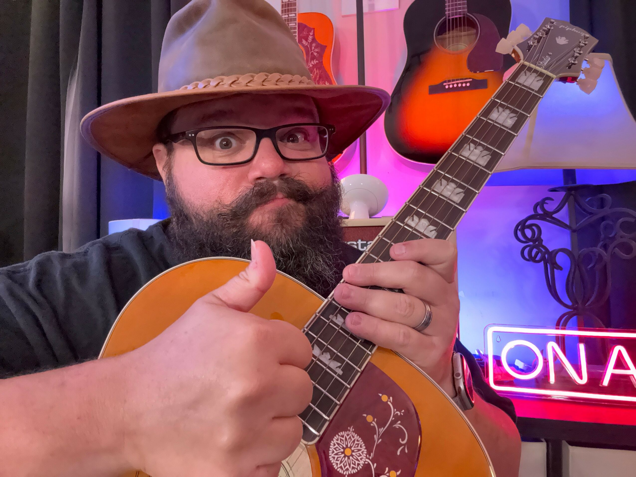 Person playing guitar, wearing glasses and a hat, upside down in a room with colorful neon lights and guitars.