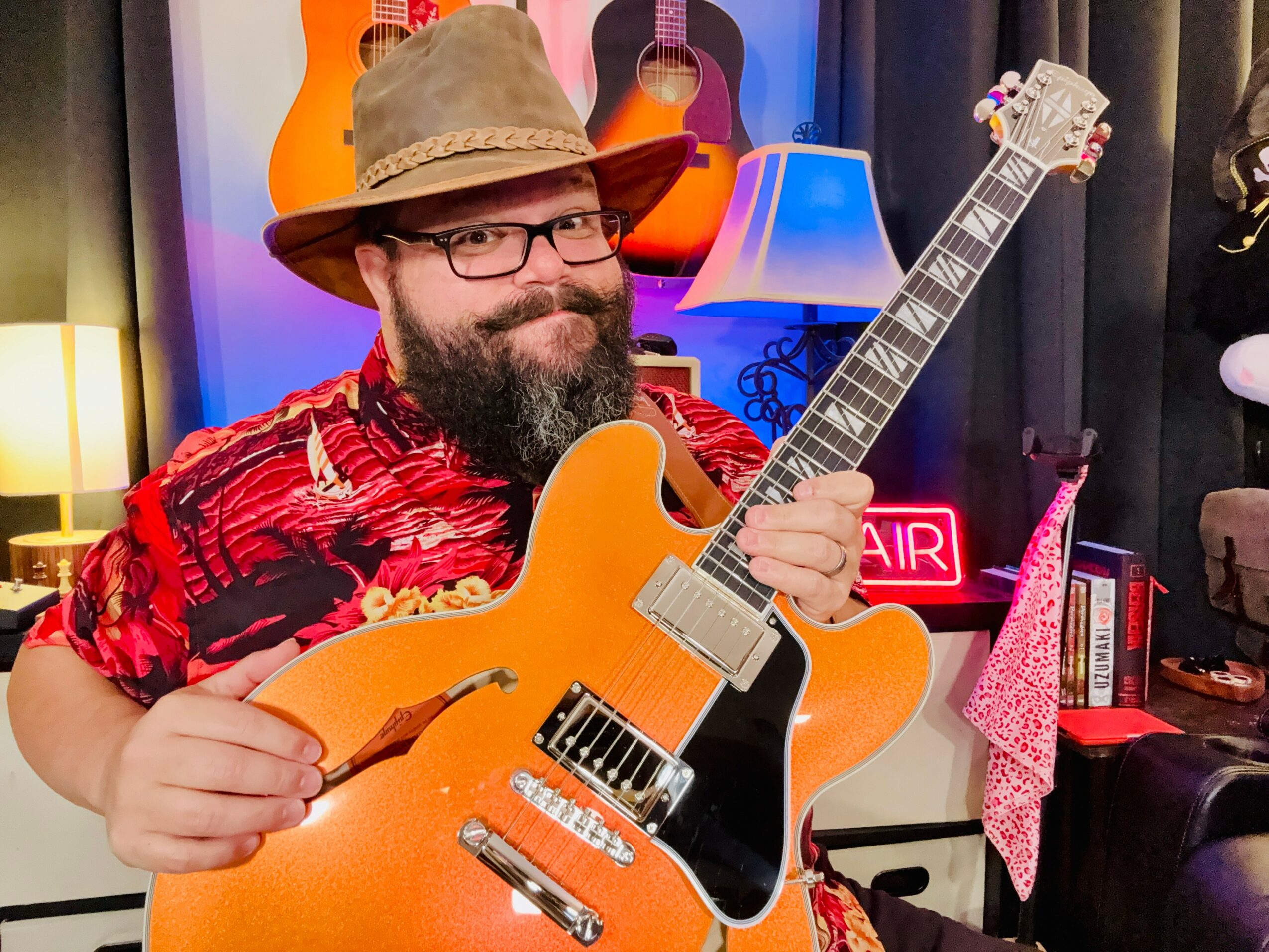 Man with glasses, beard, wearing a hat and red patterned shirt, holding an orange electric guitar in a room with guitars and books