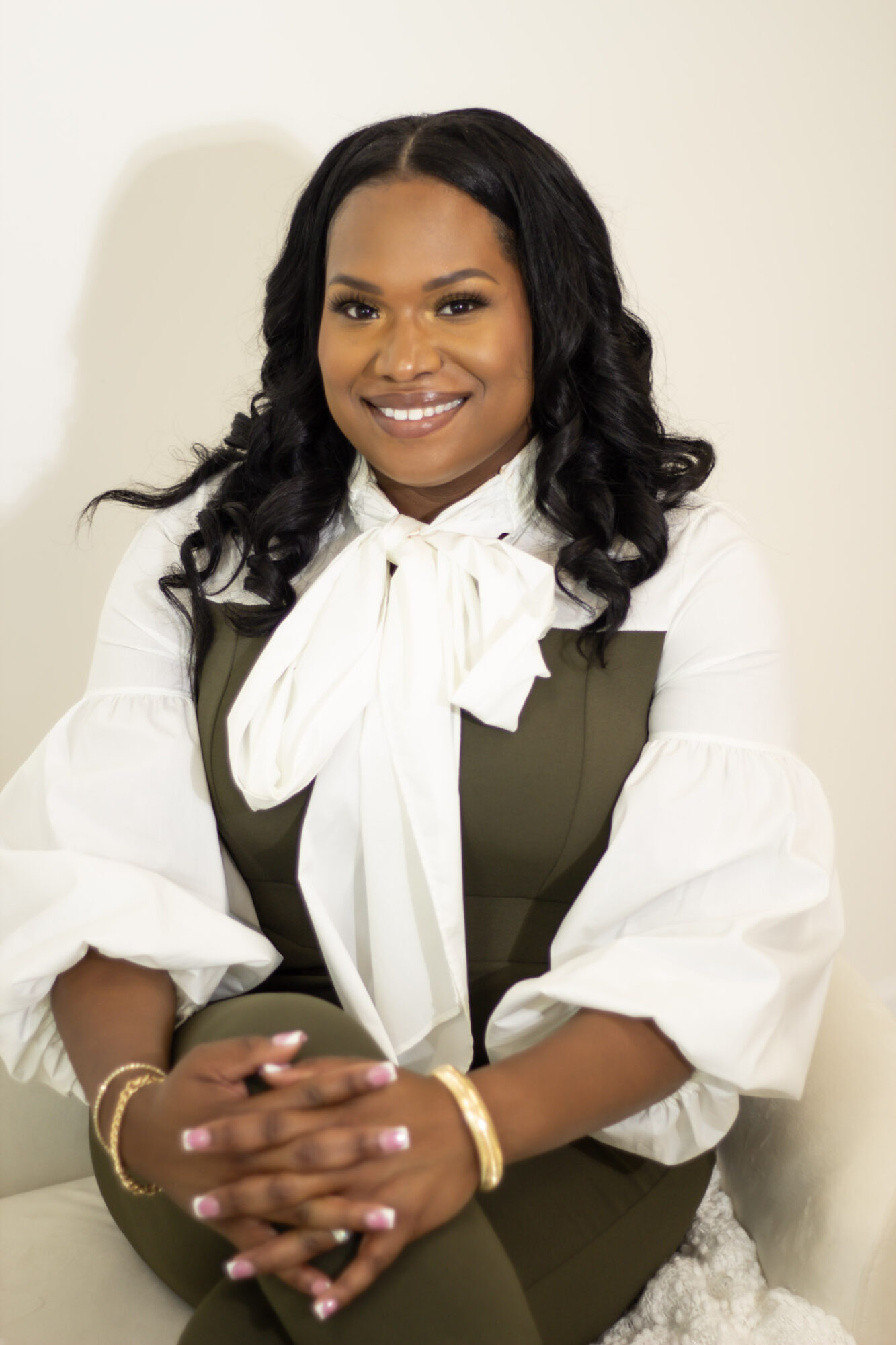 Smiling woman with long dark hair, wearing a white blouse with a bow and an olive green vest, sitting with hands clasped.