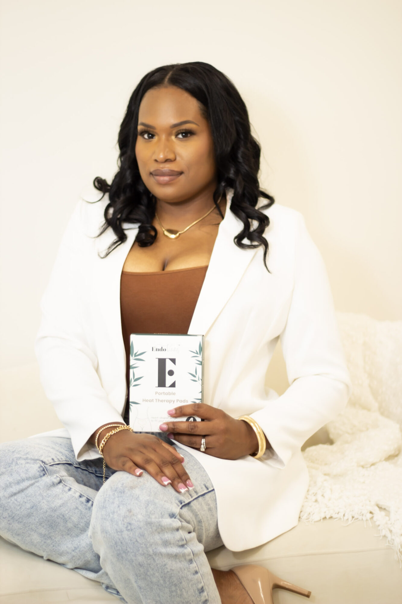 Woman with long dark hair in a white blazer holding a book, sitting on a light-colored surface, neutral background.