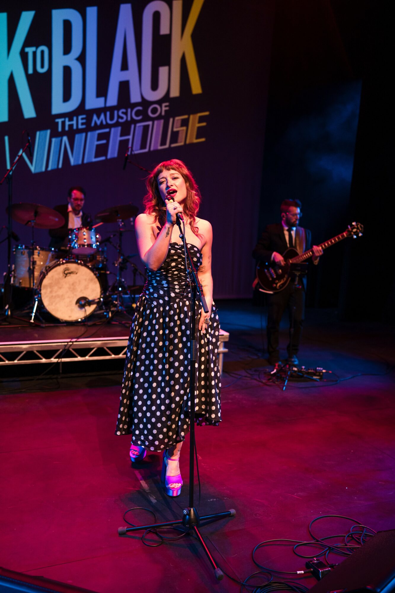 Performer singing on stage with band, colorful backdrop, and red carpeted floor.